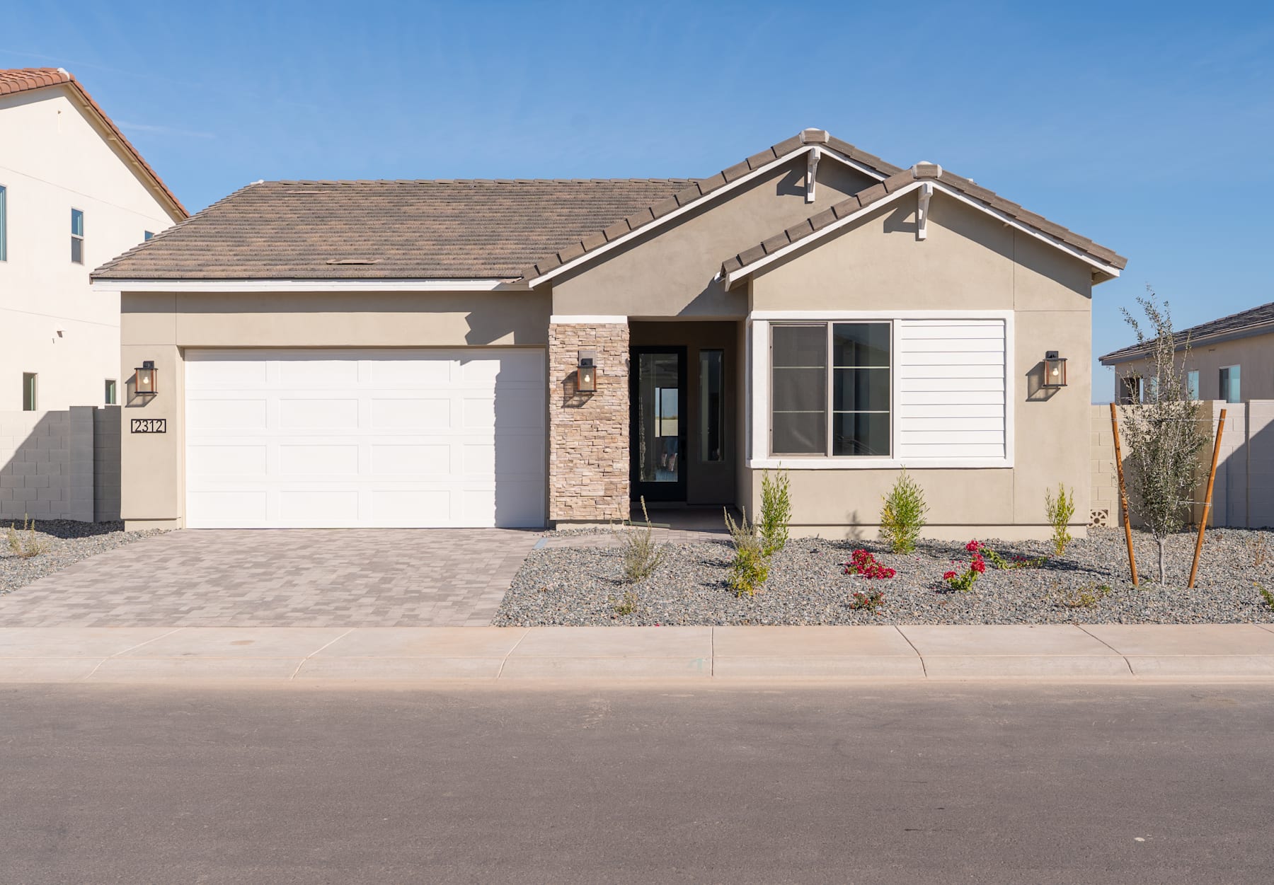 A single-story house with a garage, surrounded by a gravel driveway and landscaped with small plants, set against a clear blue sky.