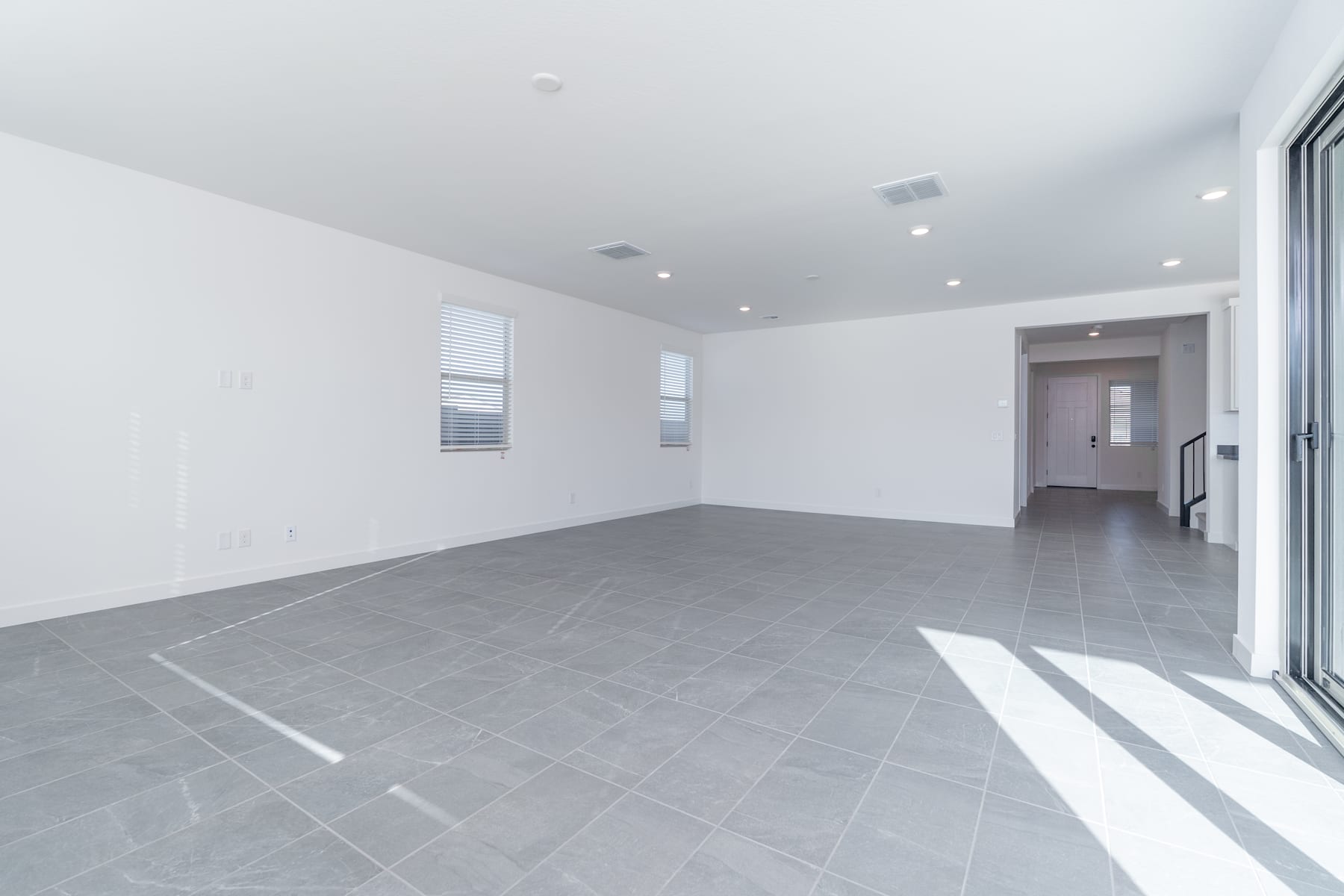 A spacious, minimalist room with white walls and a gray tiled floor, featuring a doorway leading to another area.