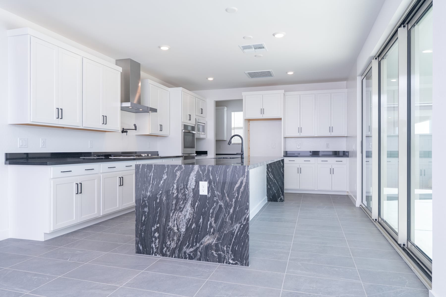 A modern, open-concept kitchen with white cabinets, a large marble island, and a sliding glass door leading to an outdoor area.