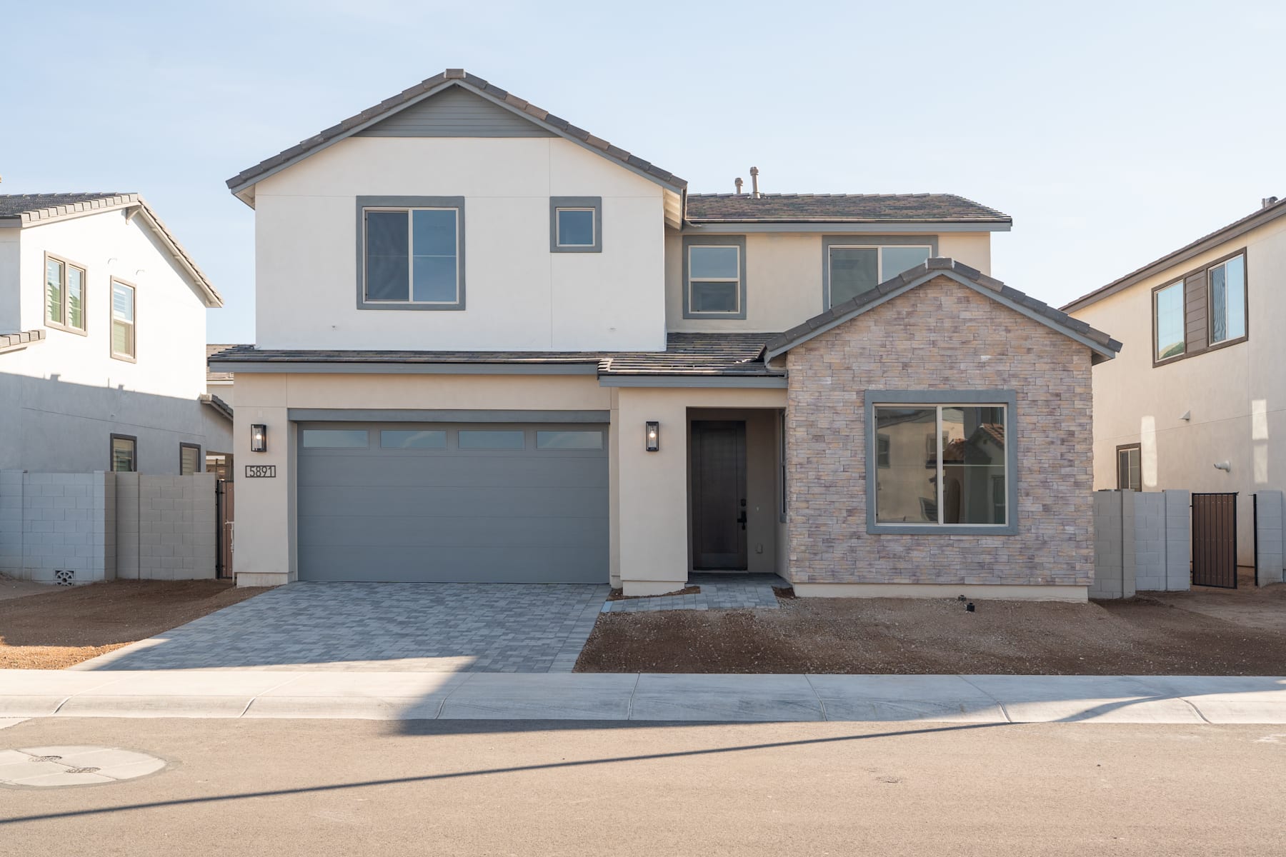 A two-story residential house with a garage, surrounded by a paved driveway and landscaped yard, set against a clear blue sky.