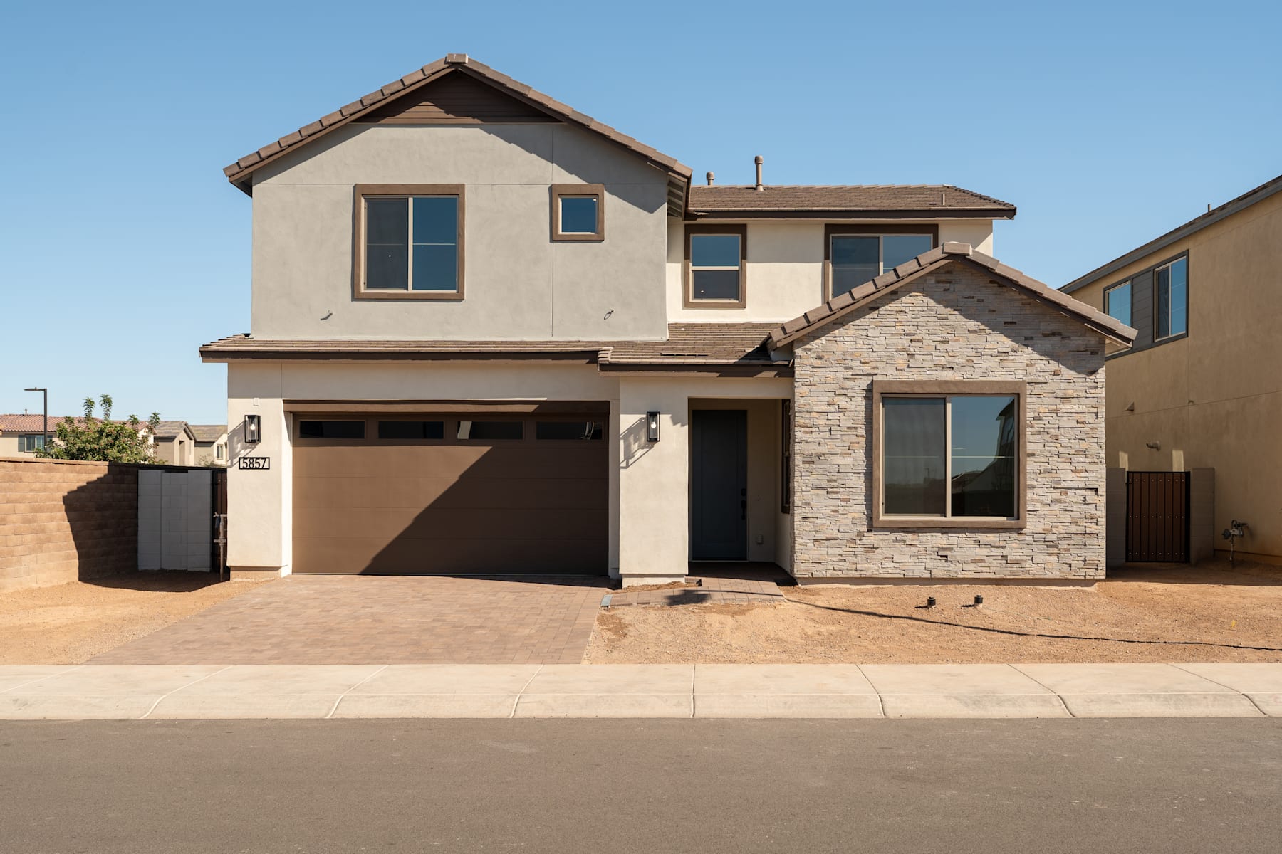A two-story residential house with a stone exterior, a garage, and a paved driveway in the foreground, set against a clear blue sky in the background.