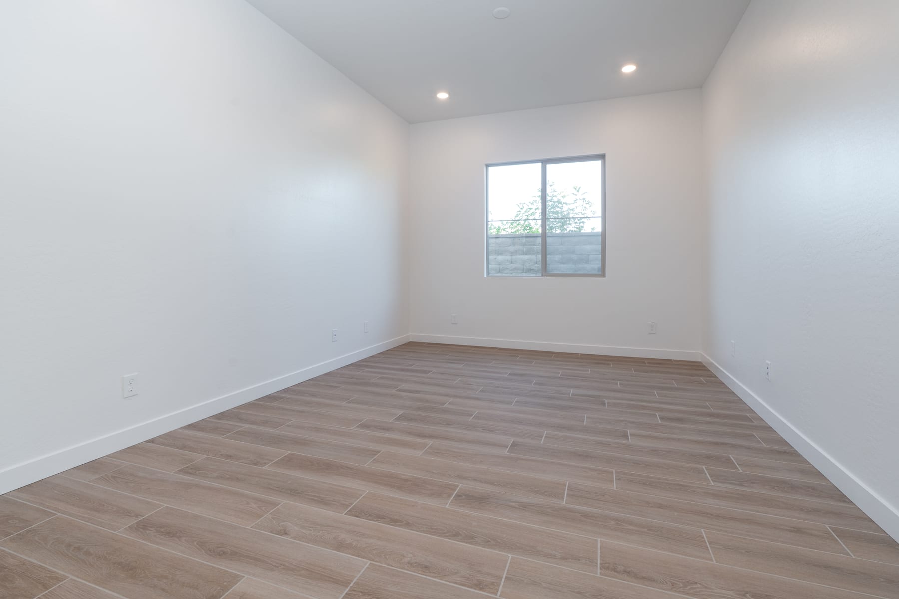A bright, empty room with white walls and a large window, featuring a wooden floor with a tile-like pattern.