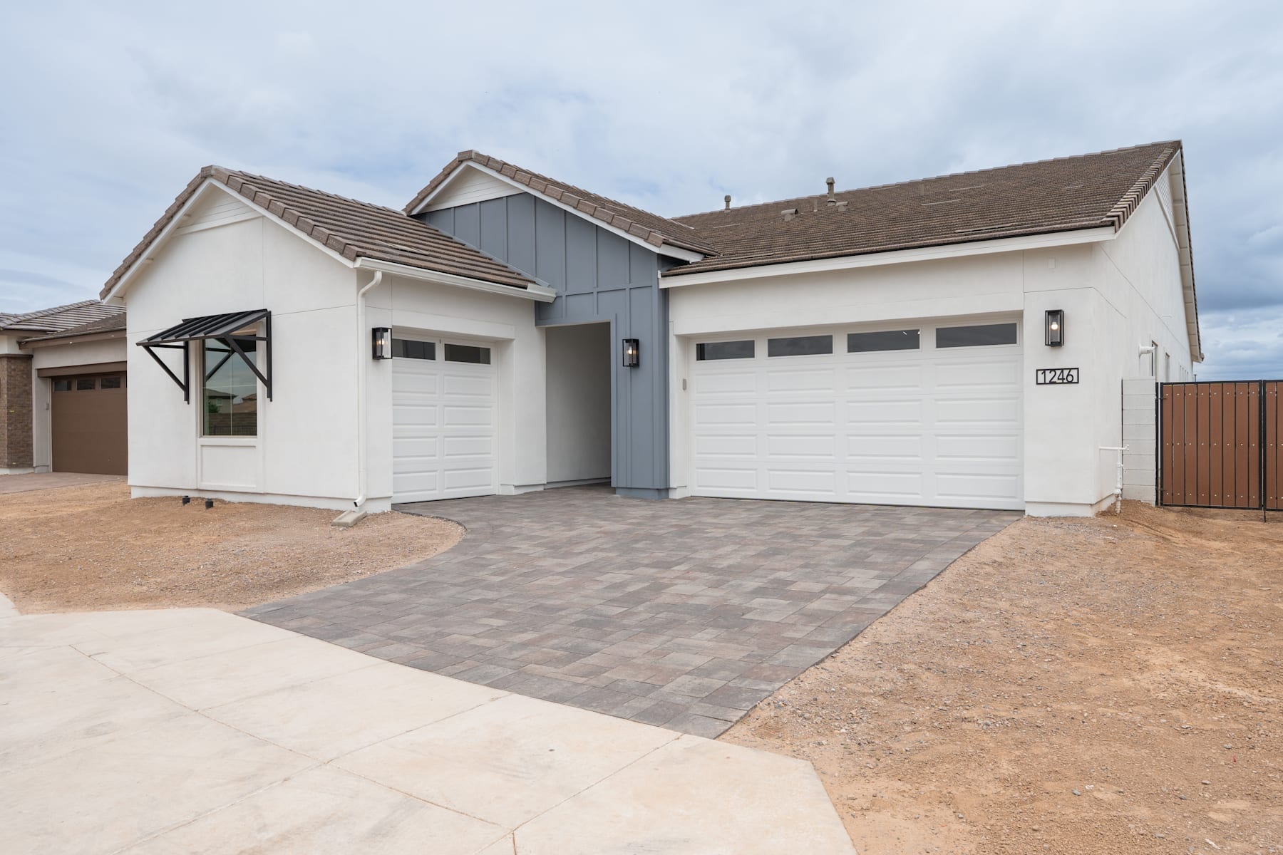 A modern, two-story house with a white exterior, gray accents, and a paved driveway leading to a pair of garage doors in the foreground. The sky in the background appears overcast.