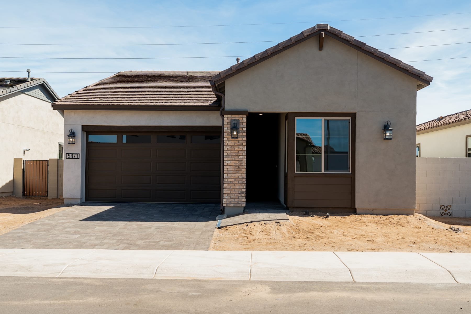 A modern, single-story house with a garage door, brick accents, and a paved driveway in the foreground, set against a clear blue sky in the background.