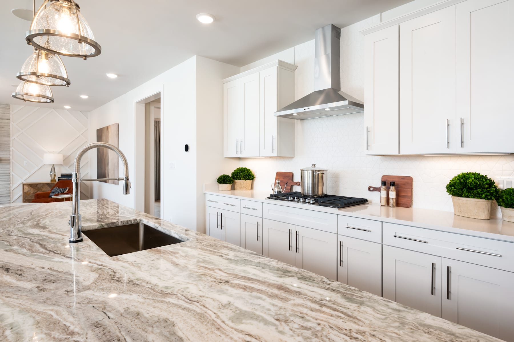 A modern, well-lit kitchen with white cabinets, a marble countertop, and a sleek stainless steel appliance in the foreground, complemented by decorative elements such as potted plants and a pendant lighting fixture in the background.