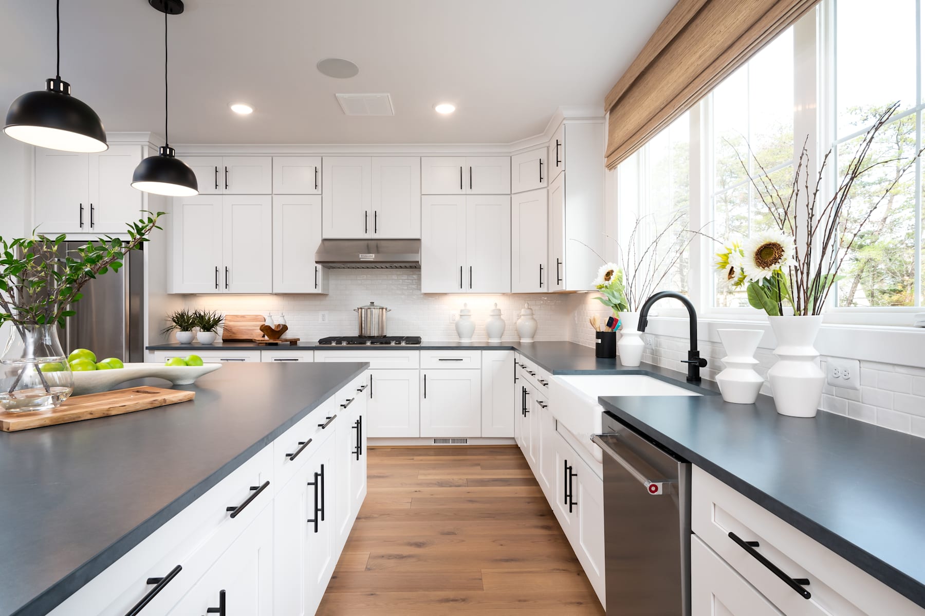 A modern, bright kitchen with white cabinets, gray countertops, and wooden floors, featuring a large window that lets in natural light and showcases a vase of sunflowers on the windowsill.