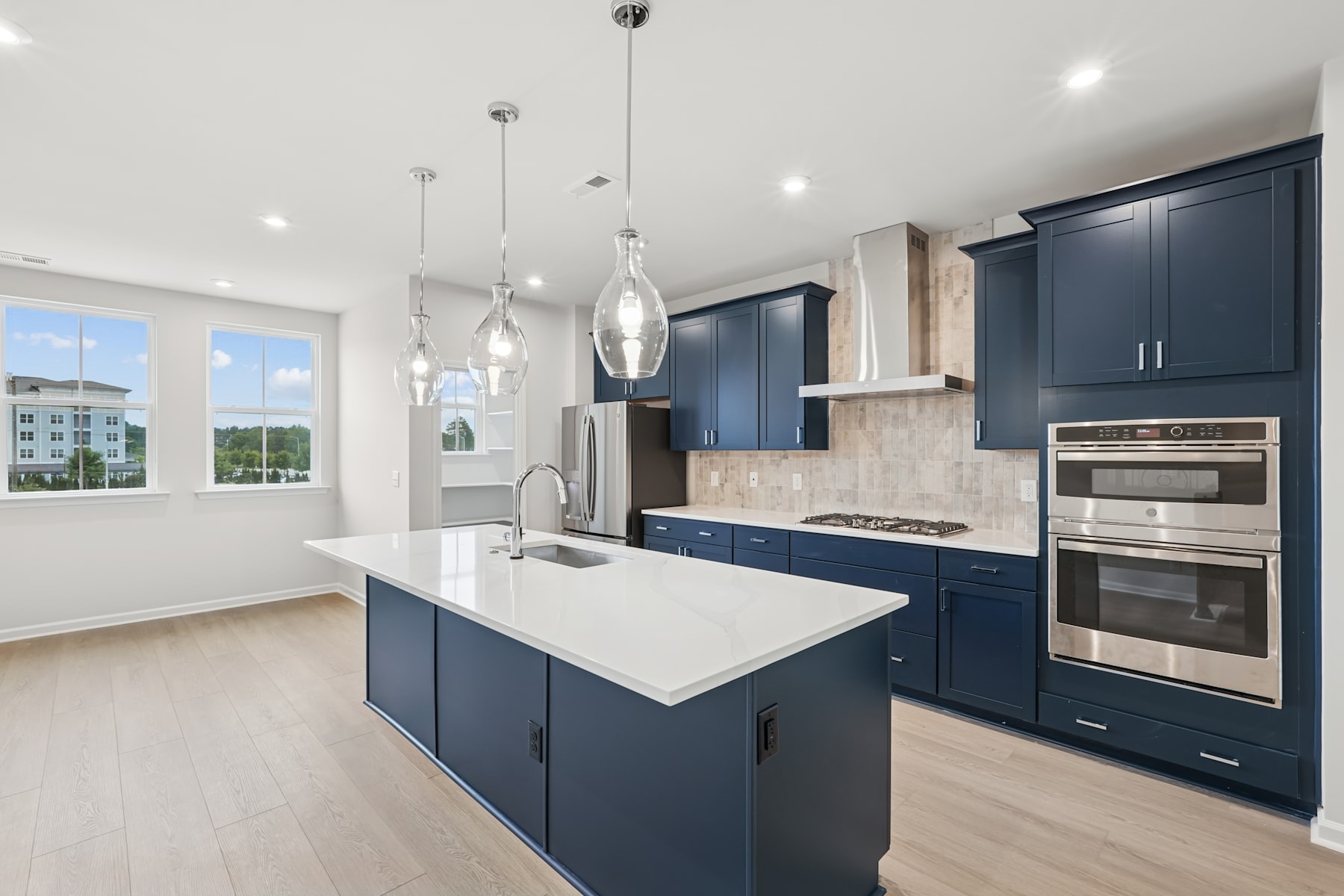 A modern, open-concept kitchen with navy blue cabinets, a white quartz countertop, and pendant lighting fixtures, set against a backdrop of large windows overlooking a scenic outdoor view.