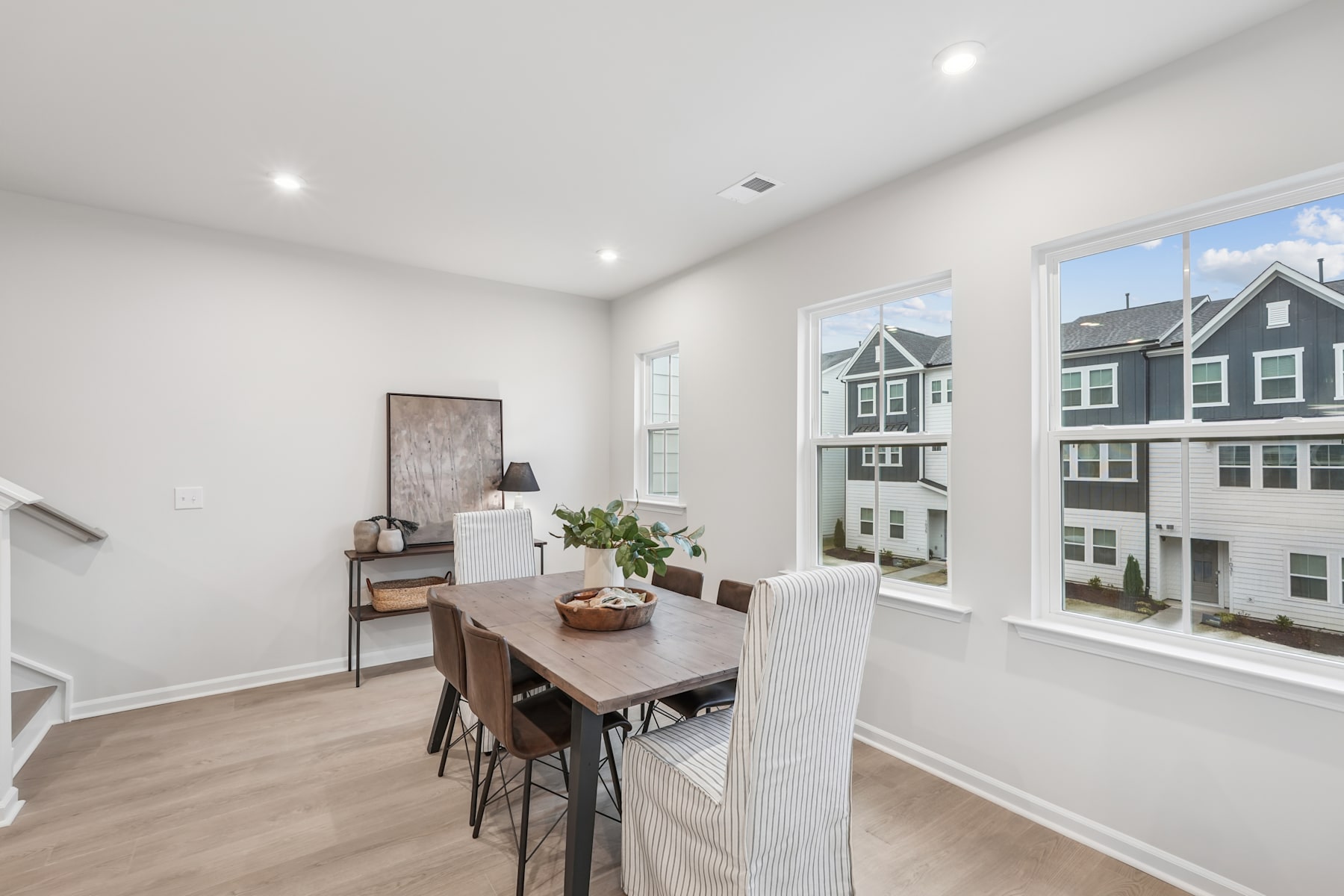 A modern, open-concept dining area with a wooden table and chairs, surrounded by large windows overlooking a residential neighborhood.