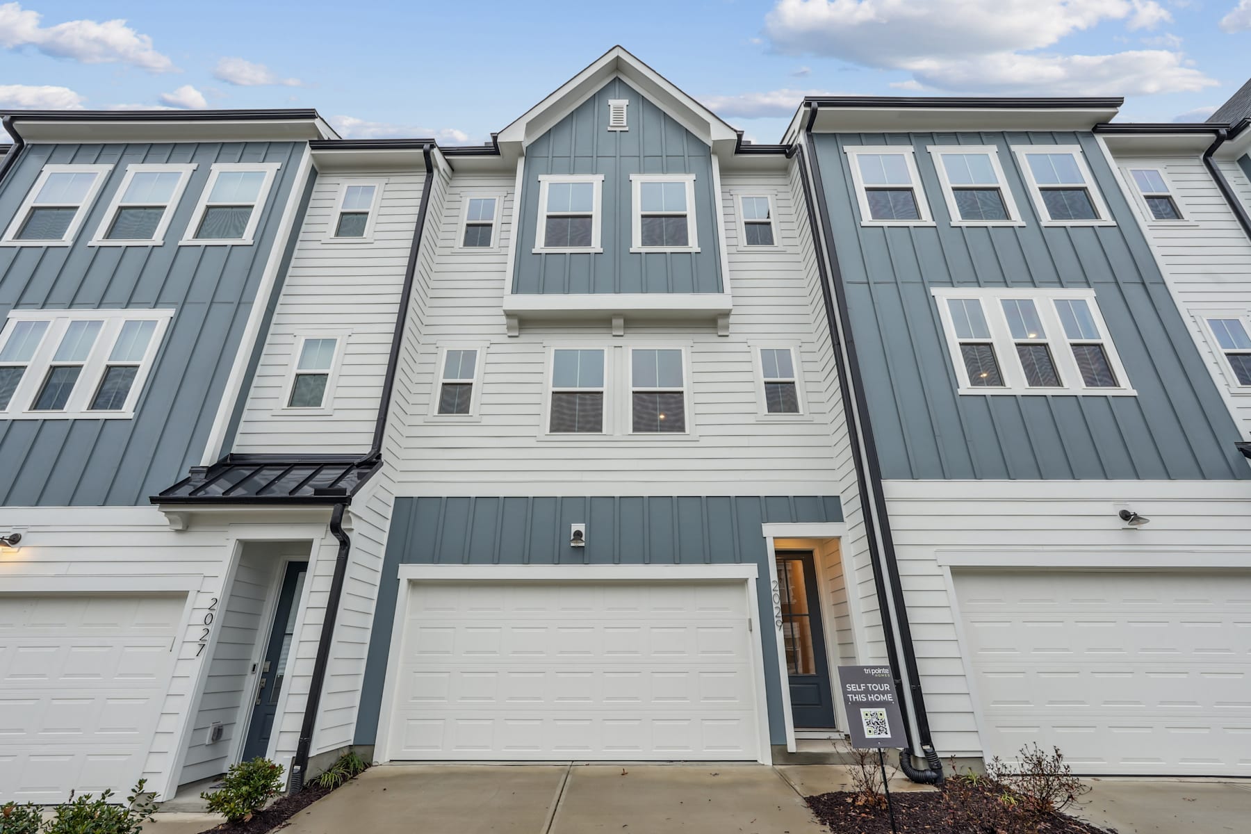 A multi-story townhouse with a gray and white exterior, featuring a garage door, windows, and a front entrance, set against a backdrop of a blue sky with scattered clouds.