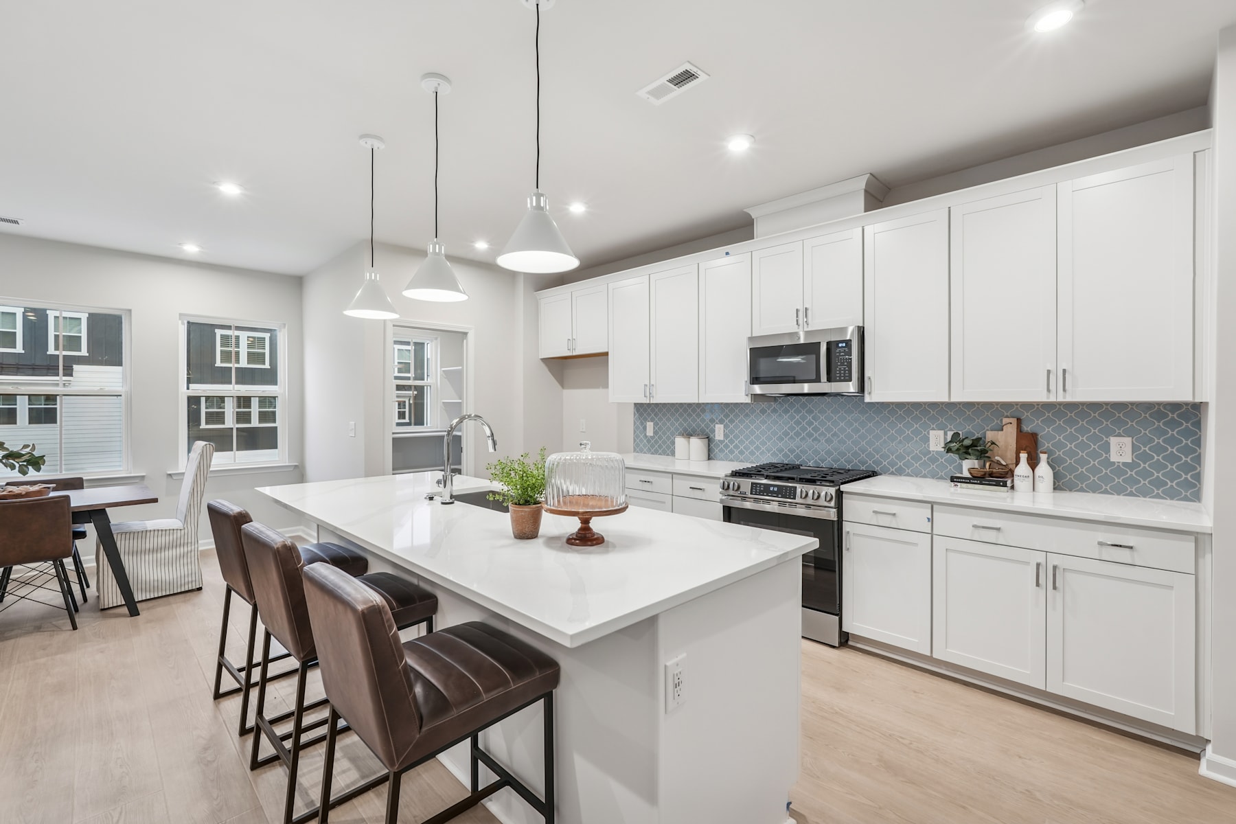 A modern, open-concept kitchen with white cabinets, a large island with bar stools, and a tiled backsplash, set against a backdrop of windows and natural light.