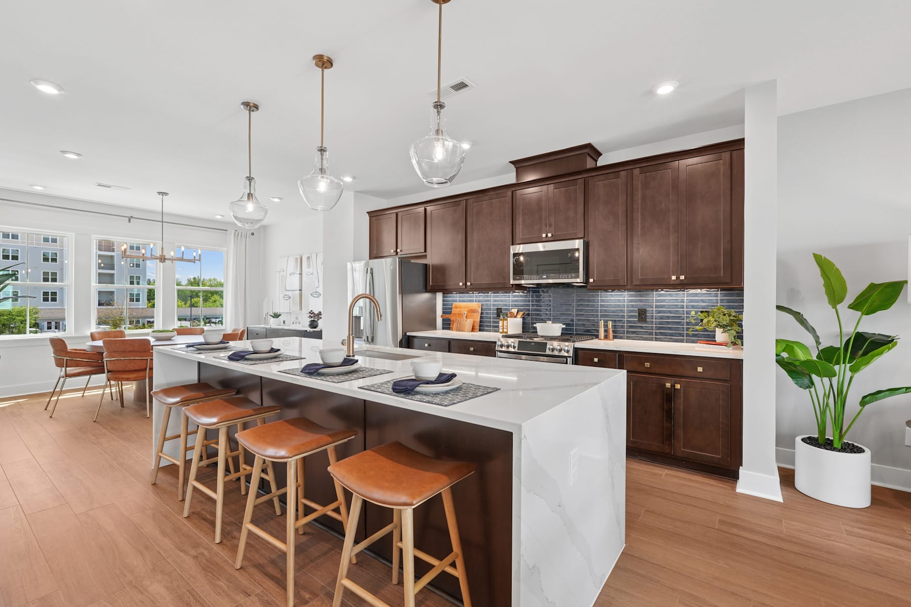 A modern and spacious kitchen with dark wood cabinets, a large island with bar stools, and pendant lighting fixtures, set against a backdrop of large windows overlooking a scenic outdoor view.
