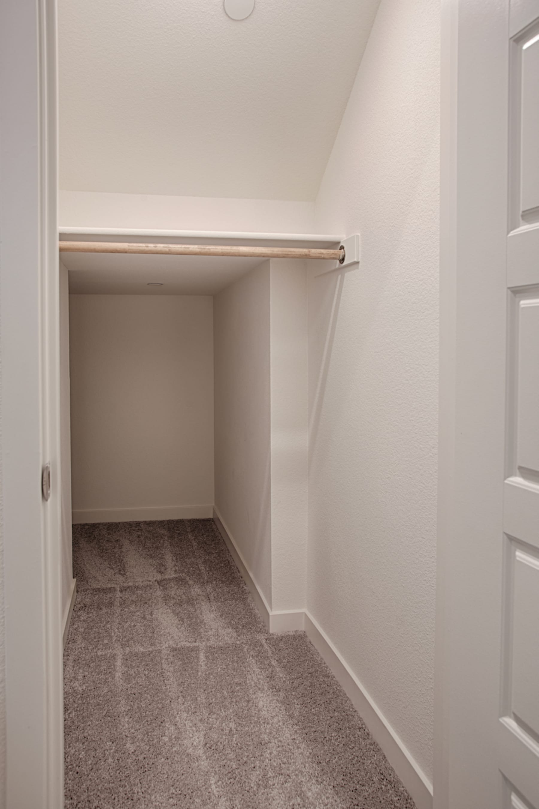 A small, empty room with white walls, a carpeted floor, and a wooden rod or shelf visible in the background.