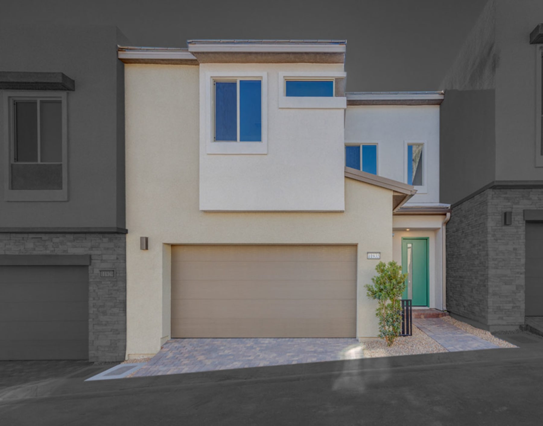 A modern, two-story residential building with a garage door, large windows, and a paved driveway in the foreground.