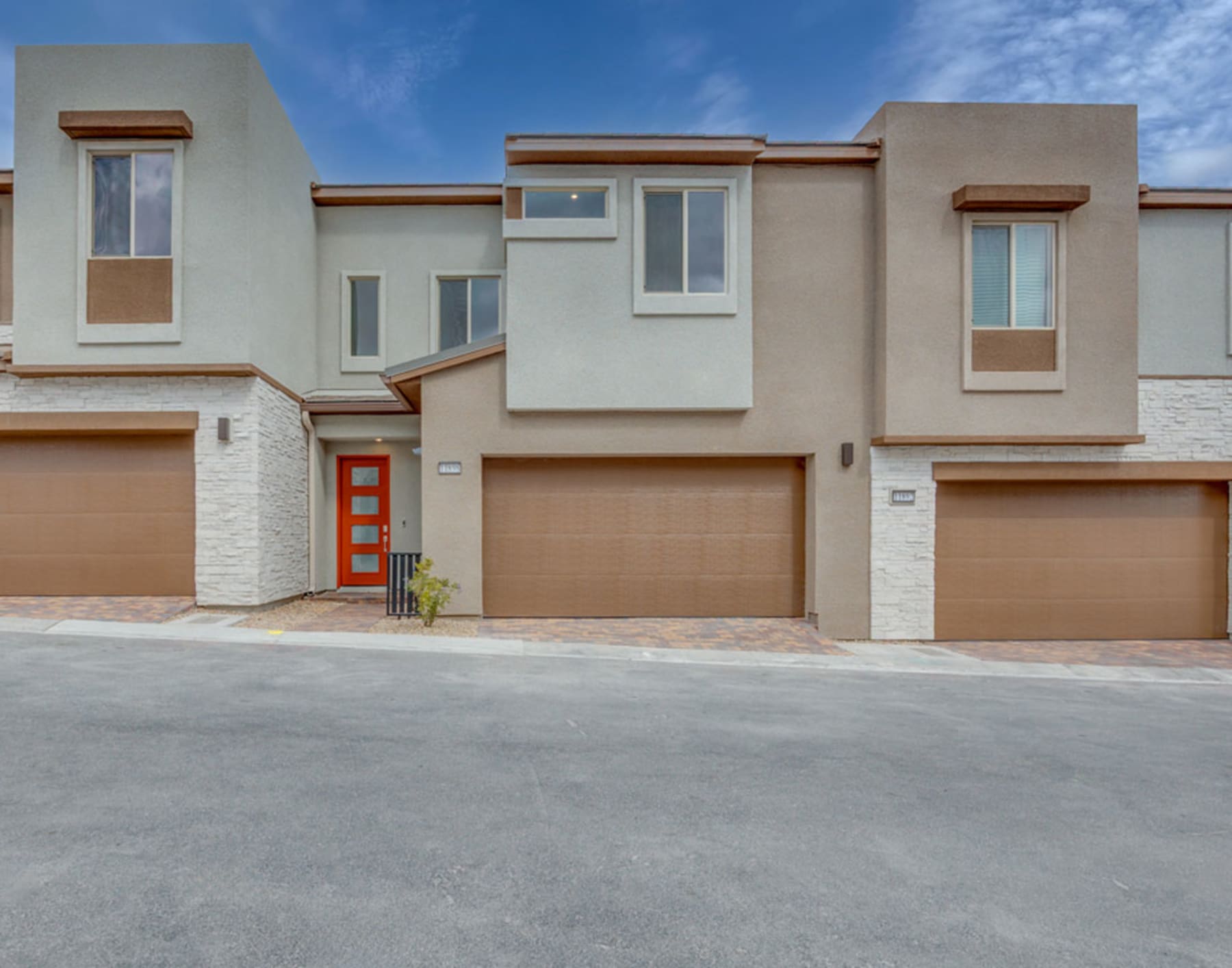The image depicts a modern, multi-unit residential building with a clean, minimalist design. The building features a mix of neutral tones, including beige and white, with accents of red around the entryway. The background shows a clear blue sky with some clouds, creating a pleasant and inviting atmosphere.