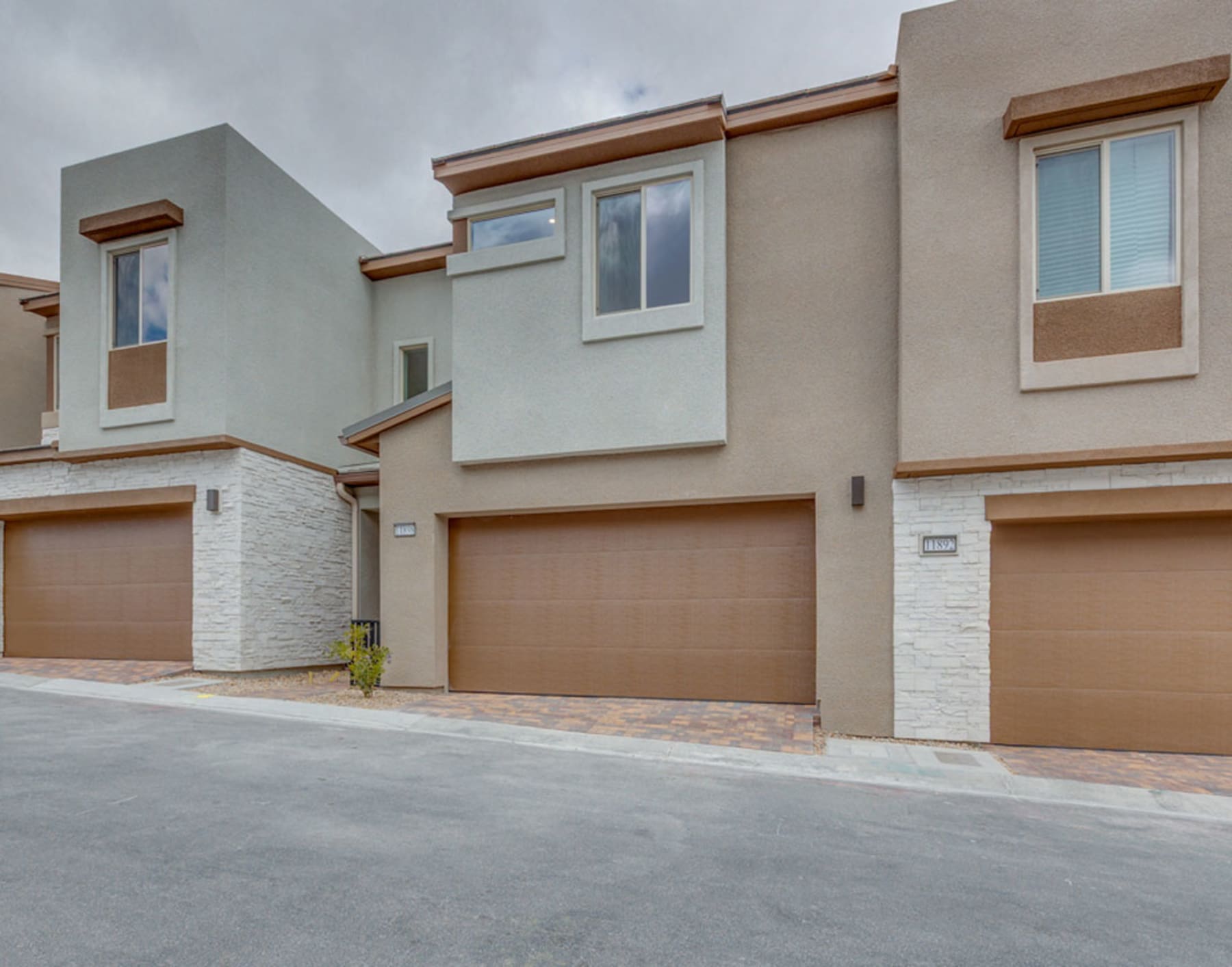 The image depicts a row of modern townhouses with beige and white exteriors, featuring wooden accents and large windows. The foreground shows a paved driveway leading up to the garages of the townhouses.