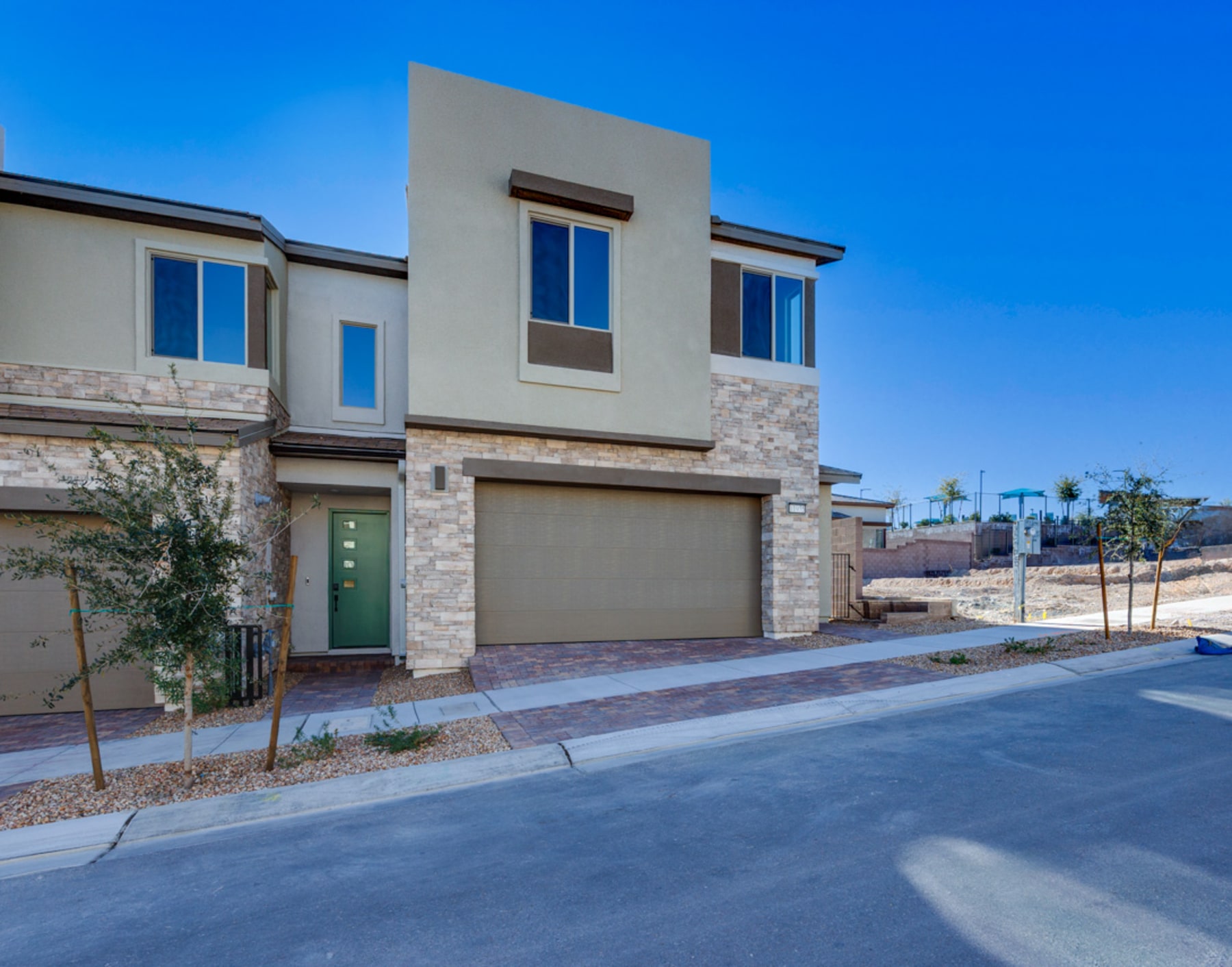 A modern, two-story townhouse with a stone facade stands in the foreground, surrounded by a paved street and landscaping in the background under a clear blue sky.