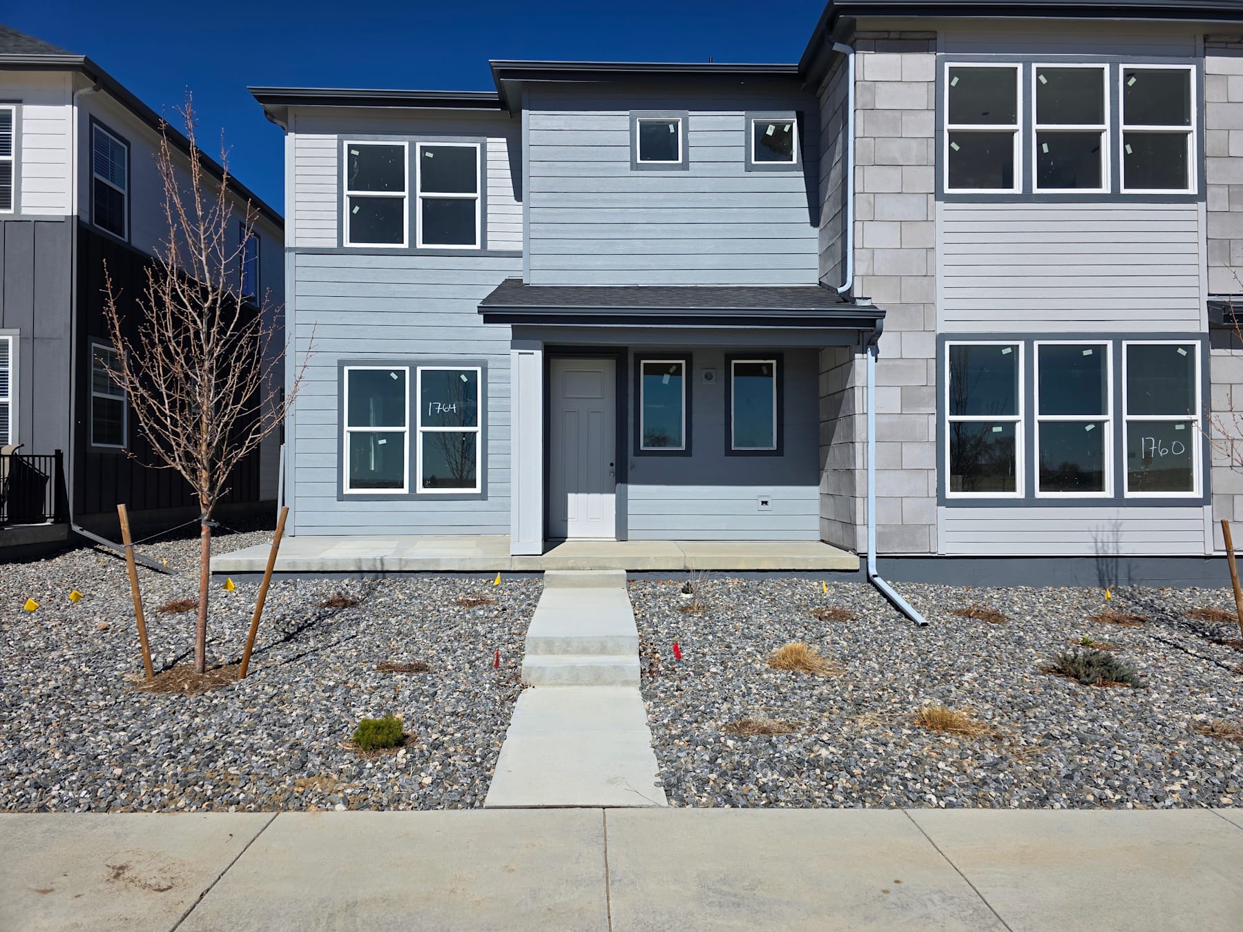 A modern, two-story residential building with a gray exterior, surrounded by a gravel pathway and landscaping, set against a clear blue sky.