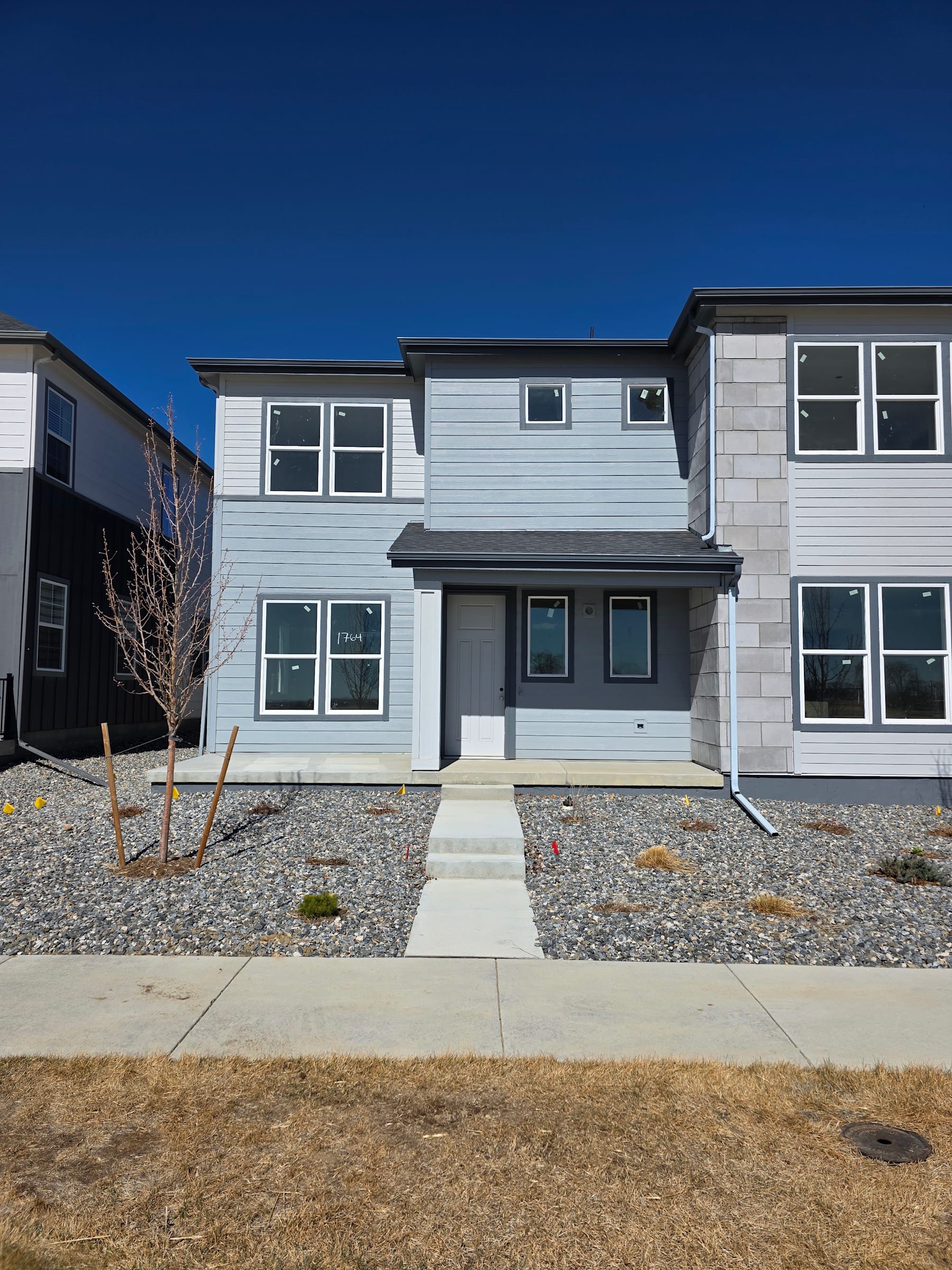 A modern, two-story residential building with a gray exterior and multiple windows, surrounded by a gravel driveway and a grassy lawn in the foreground.