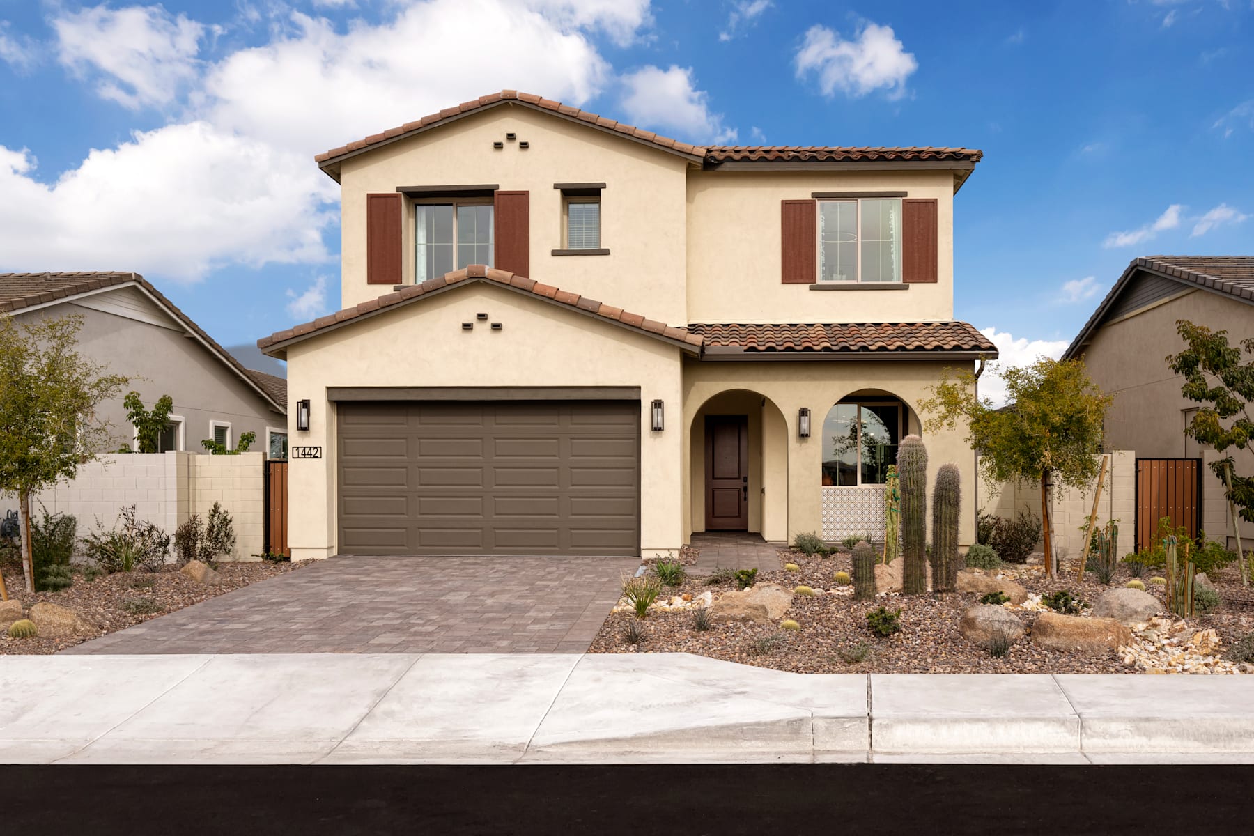 A two-story Mediterranean-style house with a tiled roof, a garage, and a well-landscaped front yard set against a blue sky with fluffy white clouds.