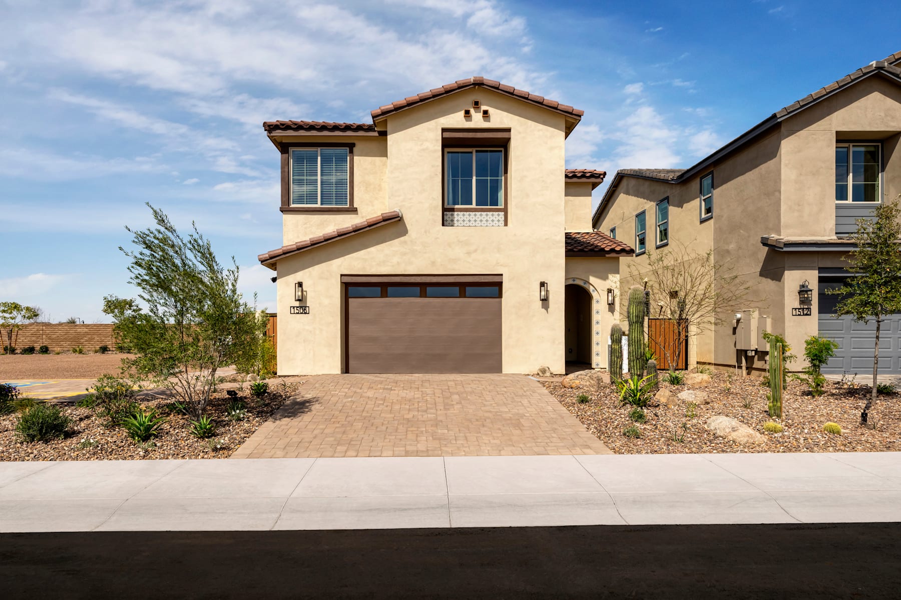 A two-story Mediterranean-style house with a tiled roof, surrounded by landscaped gardens and a paved driveway leading to a garage door.