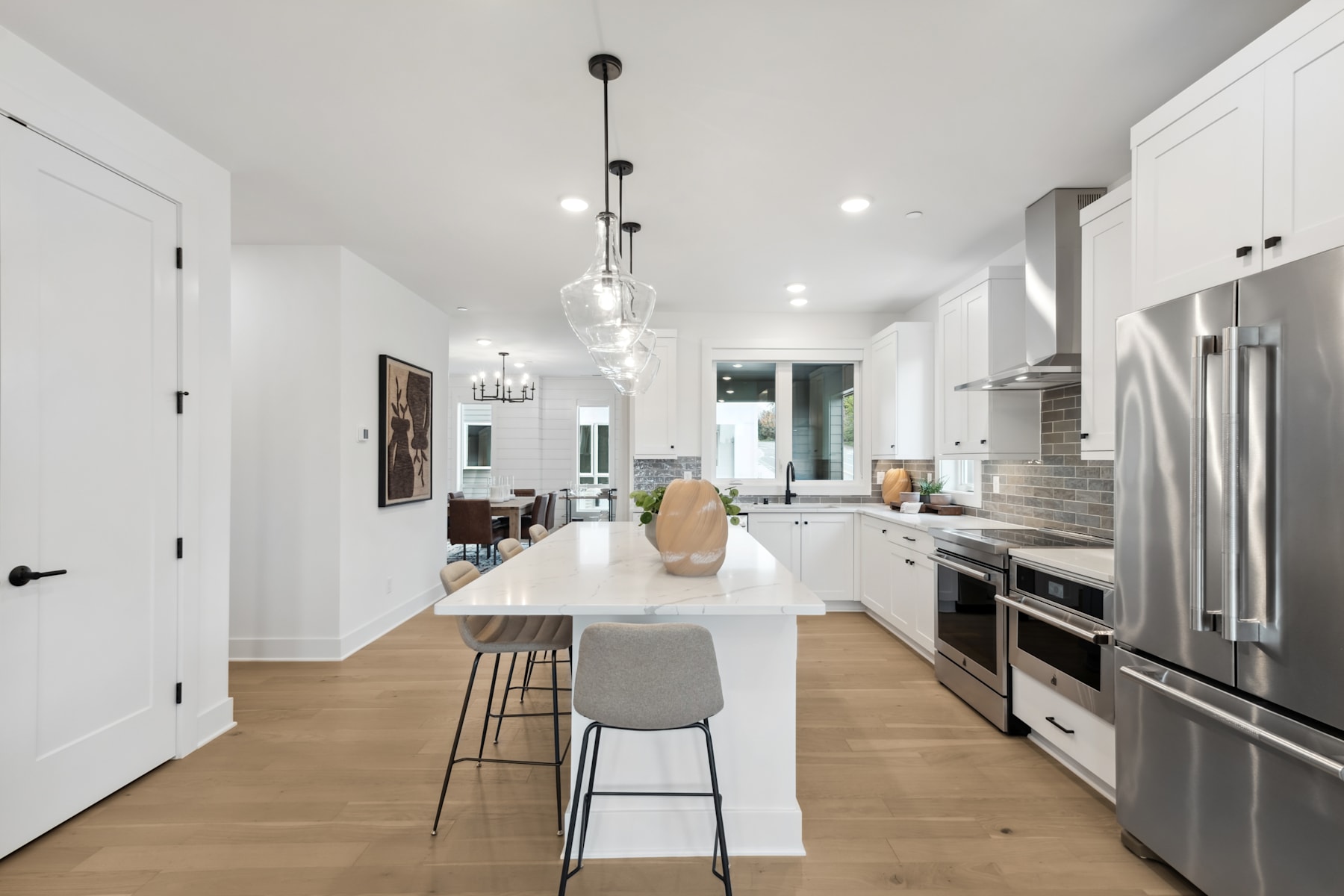 A modern, open-concept kitchen with a large white island, stainless steel appliances, and pendant lighting fixtures, set against a backdrop of white walls and hardwood floors.