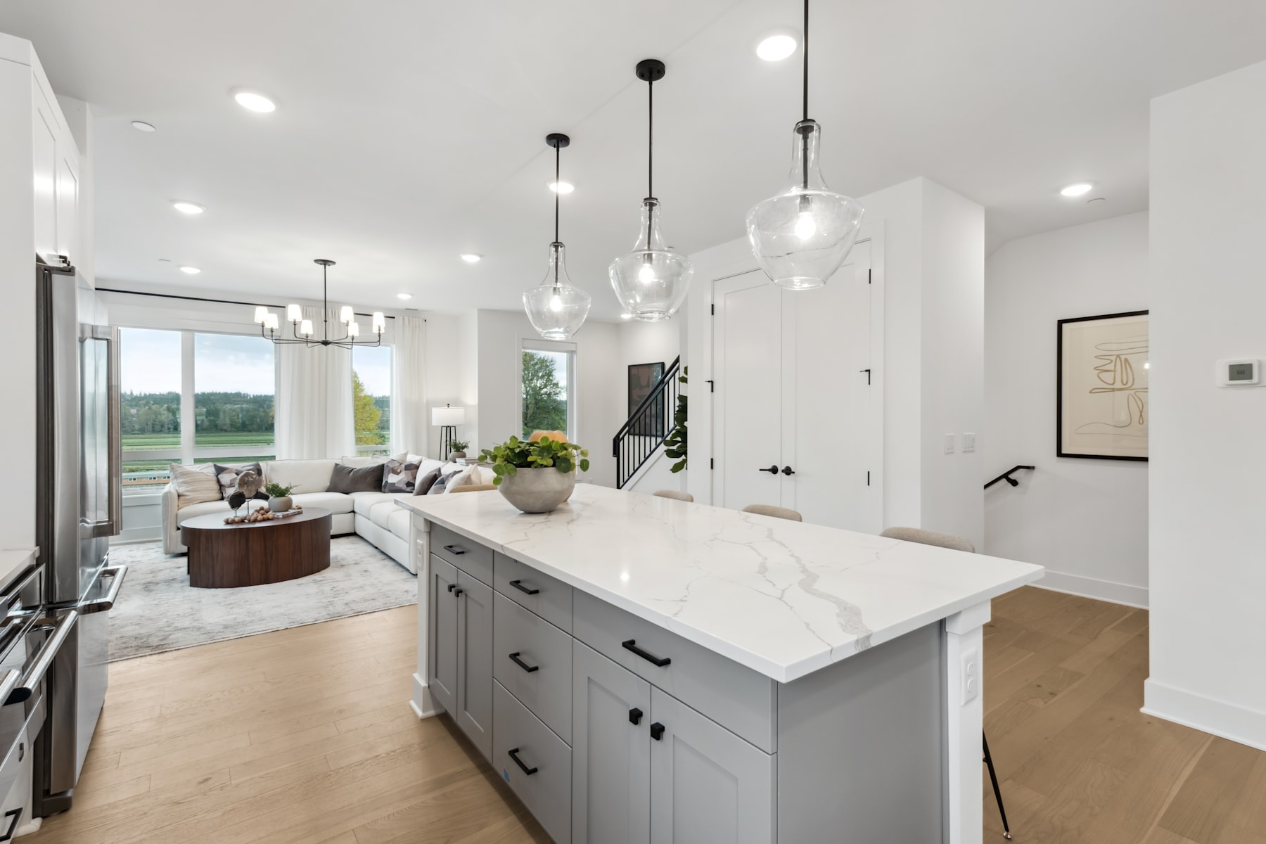 A modern, open-concept kitchen with a large island, white countertops, and pendant lighting, with a living room visible in the background.