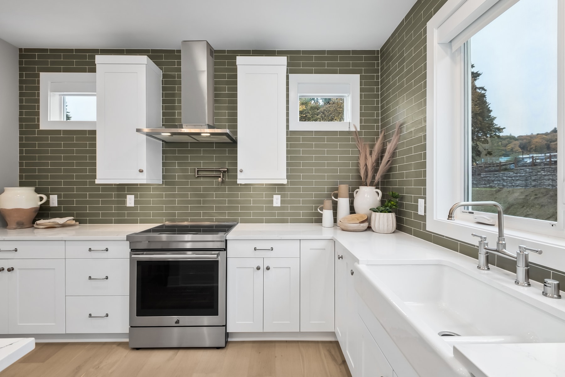 A modern, well-equipped kitchen with white cabinets, stainless steel appliances, and a backsplash of olive-green tiles, set against a scenic outdoor view through the window.