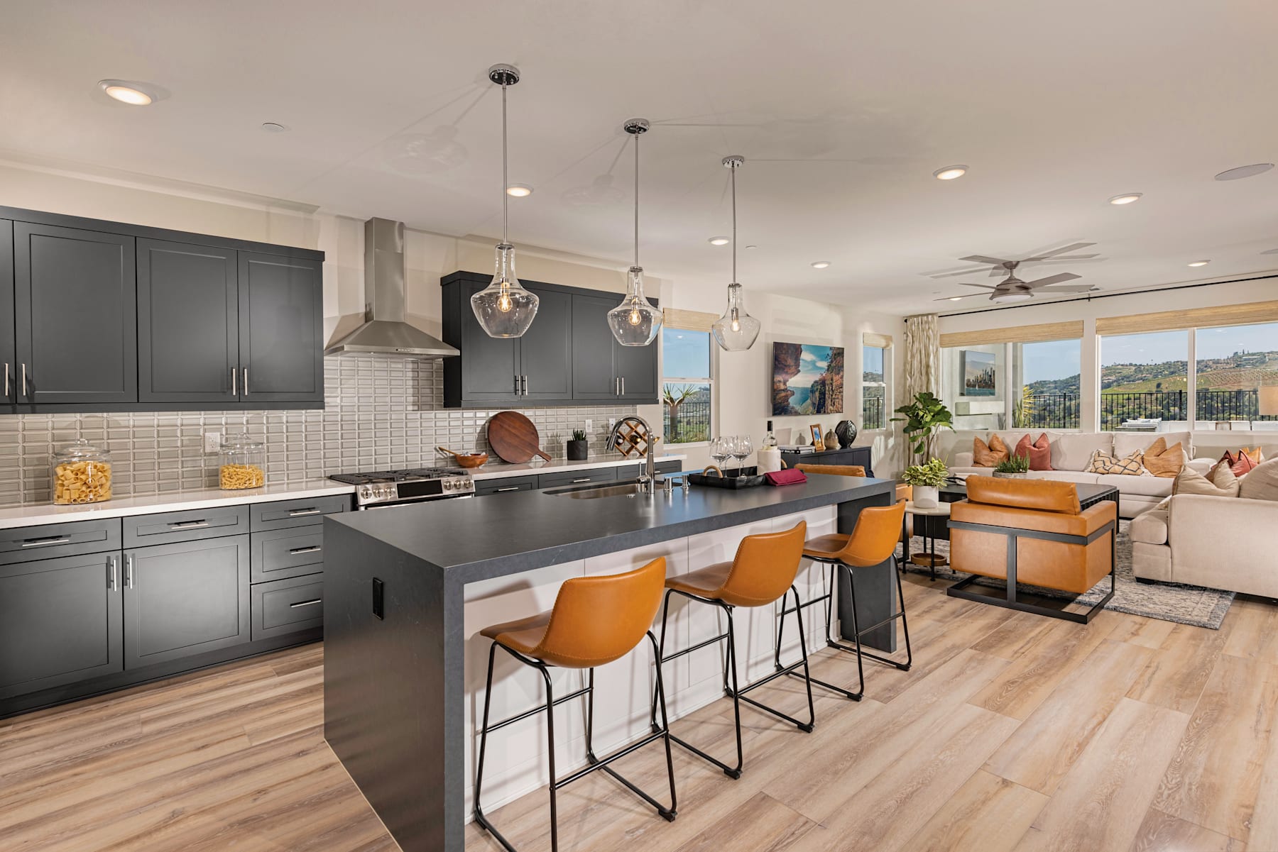A modern, open-concept kitchen and living room with sleek gray cabinets, pendant lighting, and orange bar stools, set against a backdrop of large windows overlooking a scenic outdoor view.