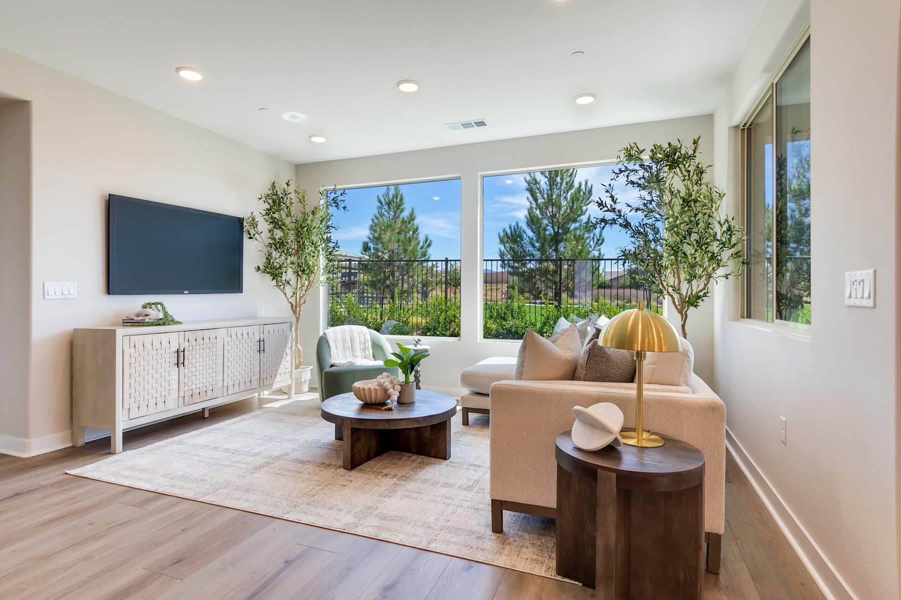 A cozy and bright living room with large windows overlooking a lush outdoor landscape, featuring a TV, a wooden coffee table, and comfortable seating arrangements.