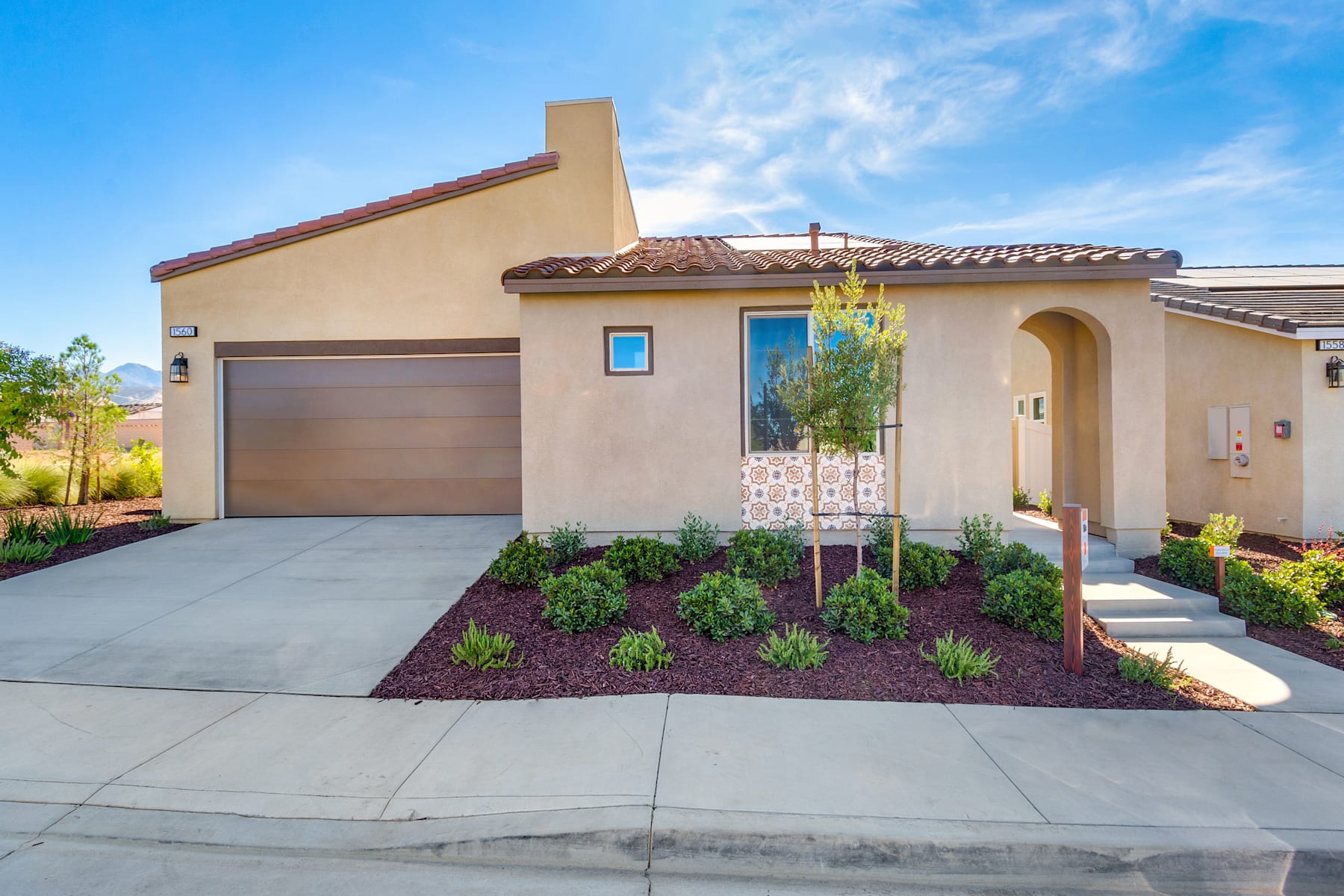 A modern, single-story house with a tiled roof, a garage door, and a well-landscaped front yard featuring various plants and a concrete walkway leading to the entrance.
