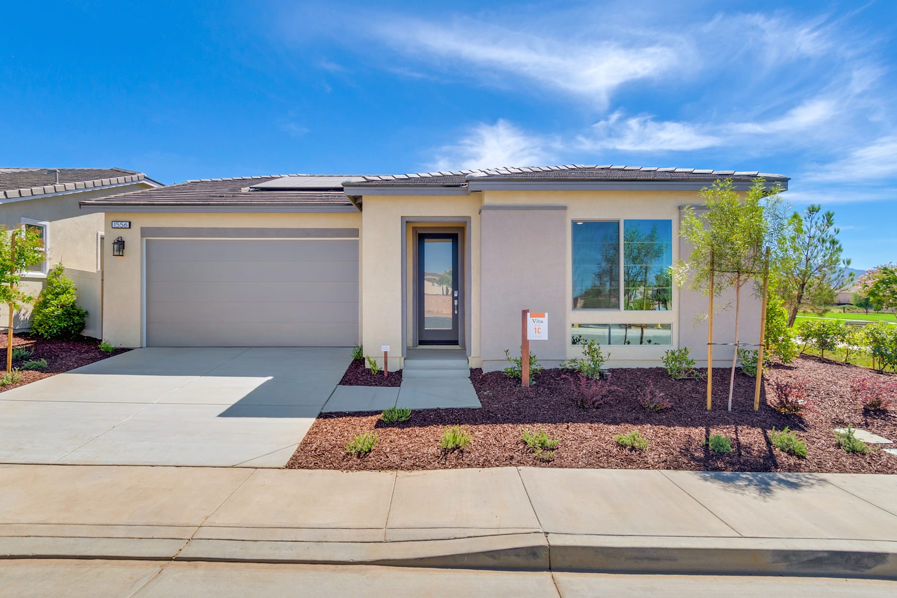 A modern, single-story house with a gray roof and beige exterior, surrounded by landscaped gardens and a paved driveway, set against a clear blue sky with wispy clouds.