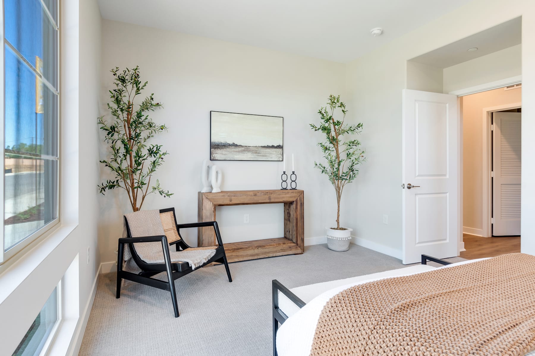 A cozy and minimalist bedroom with a wooden console, potted plants, and a framed artwork on the wall, complemented by a large window that allows natural light to fill the space.