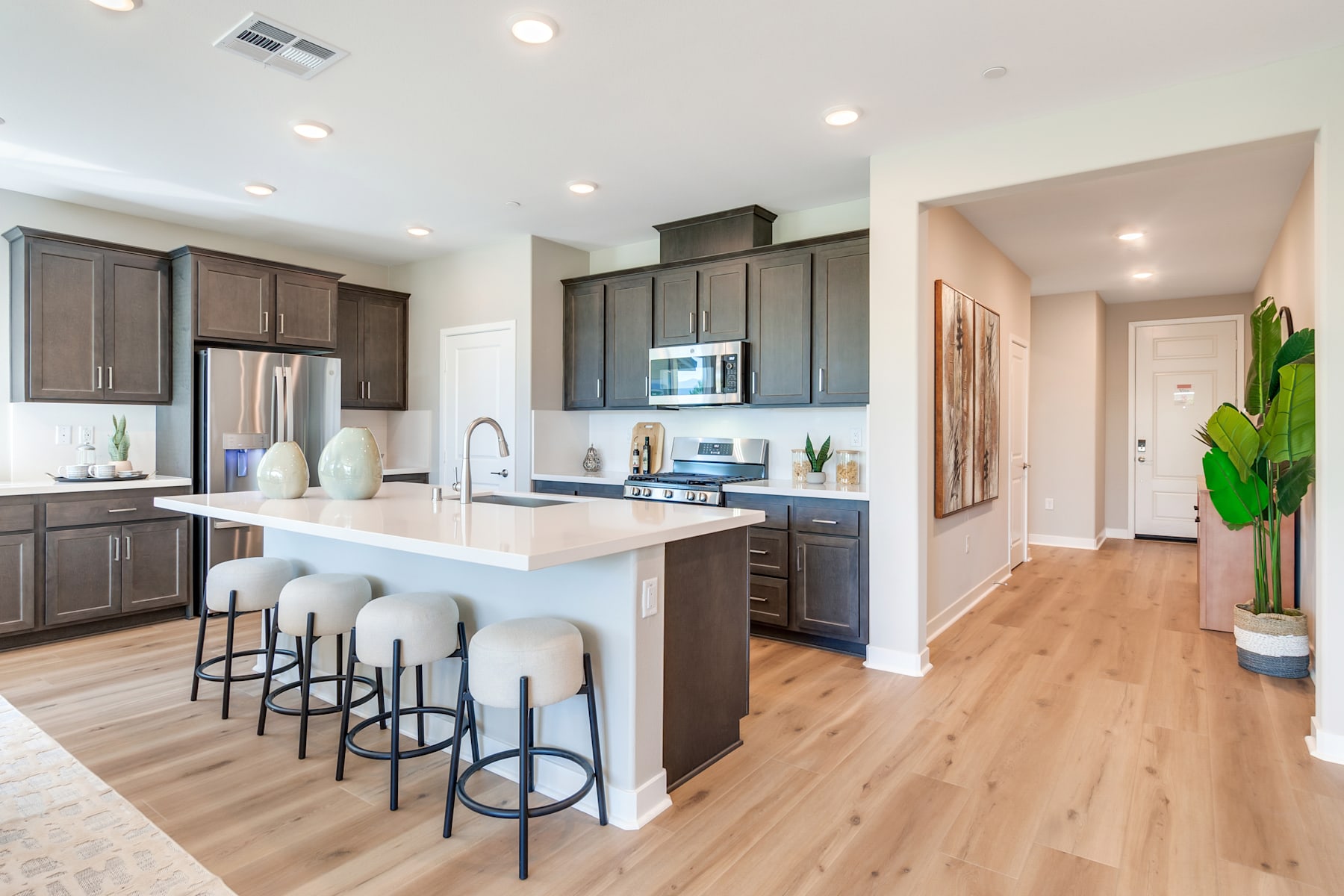 A modern and spacious kitchen with dark wood cabinets, white countertops, and bar stools, leading into a hallway with a potted plant.