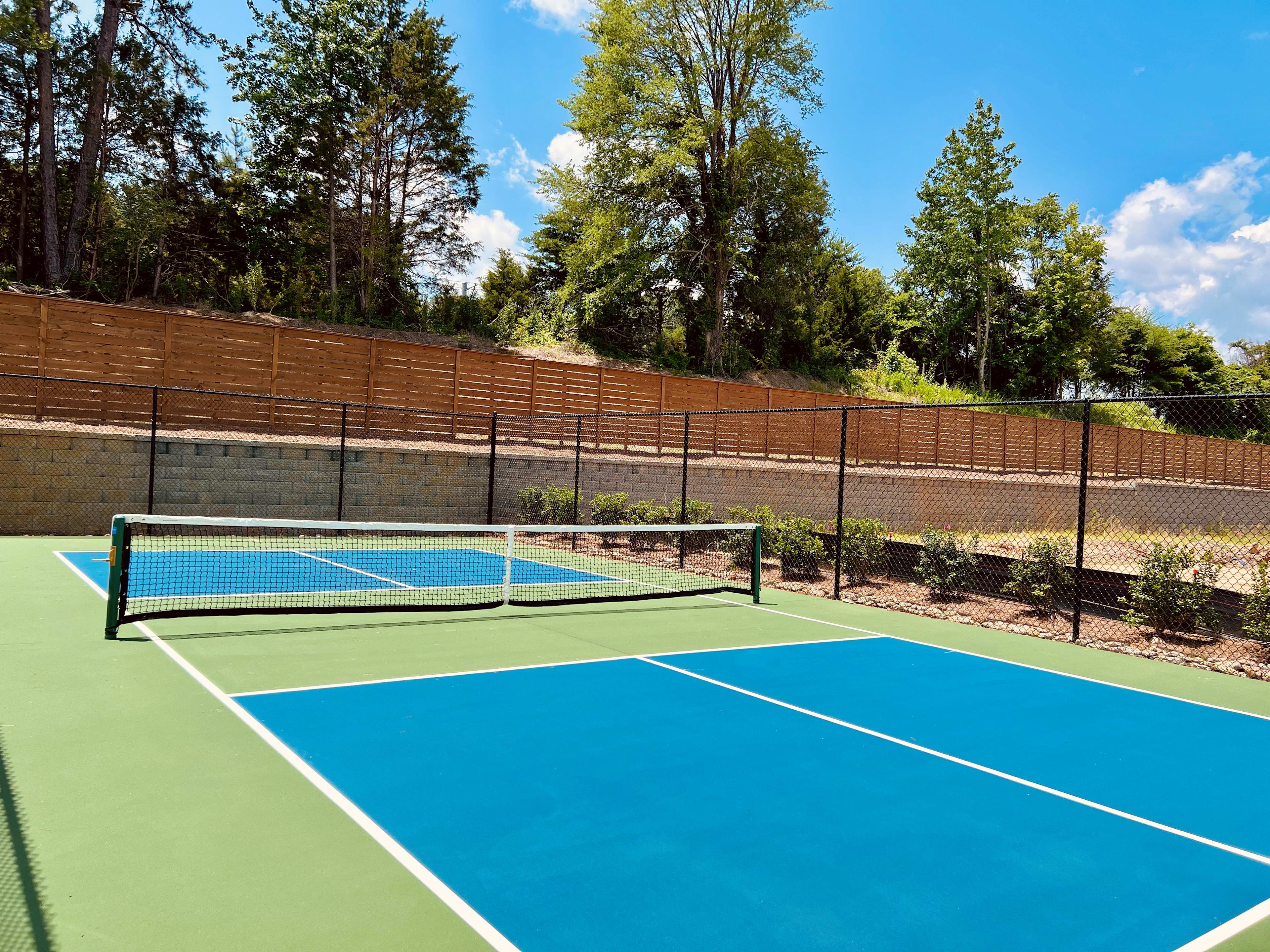 A tennis court with a blue and green surface surrounded by a wooden fence, with a forested area in the background.