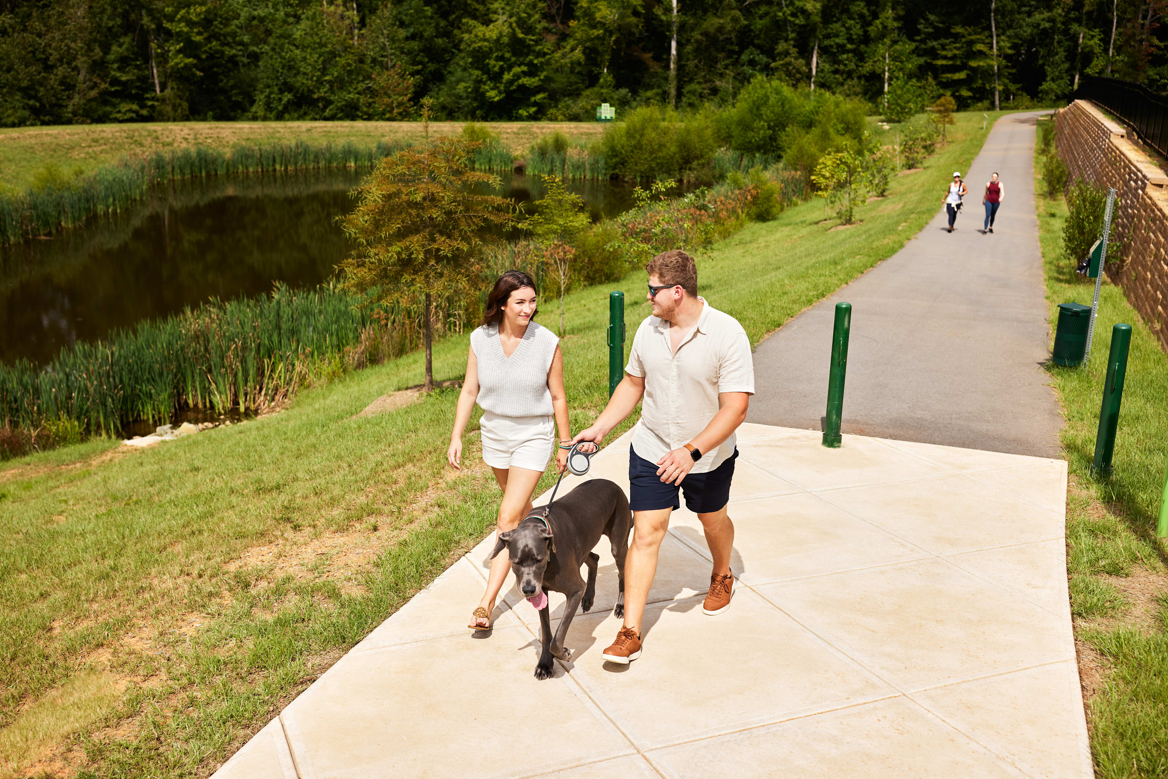 A couple walks their dog along a paved path surrounded by lush greenery, with a pond visible in the background.