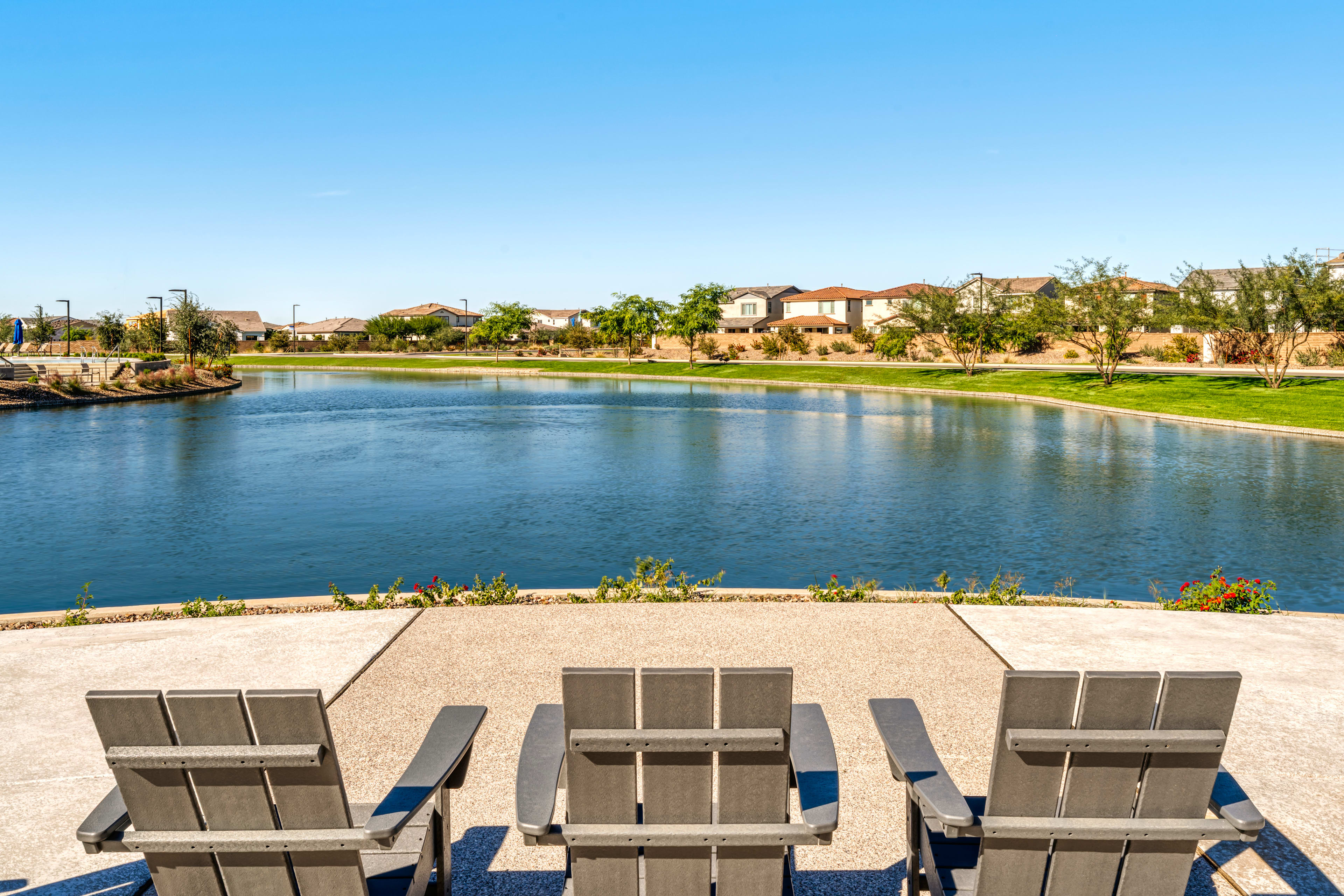 A serene lake surrounded by lush greenery and residential buildings, with three modern outdoor chairs in the foreground overlooking the tranquil scene.