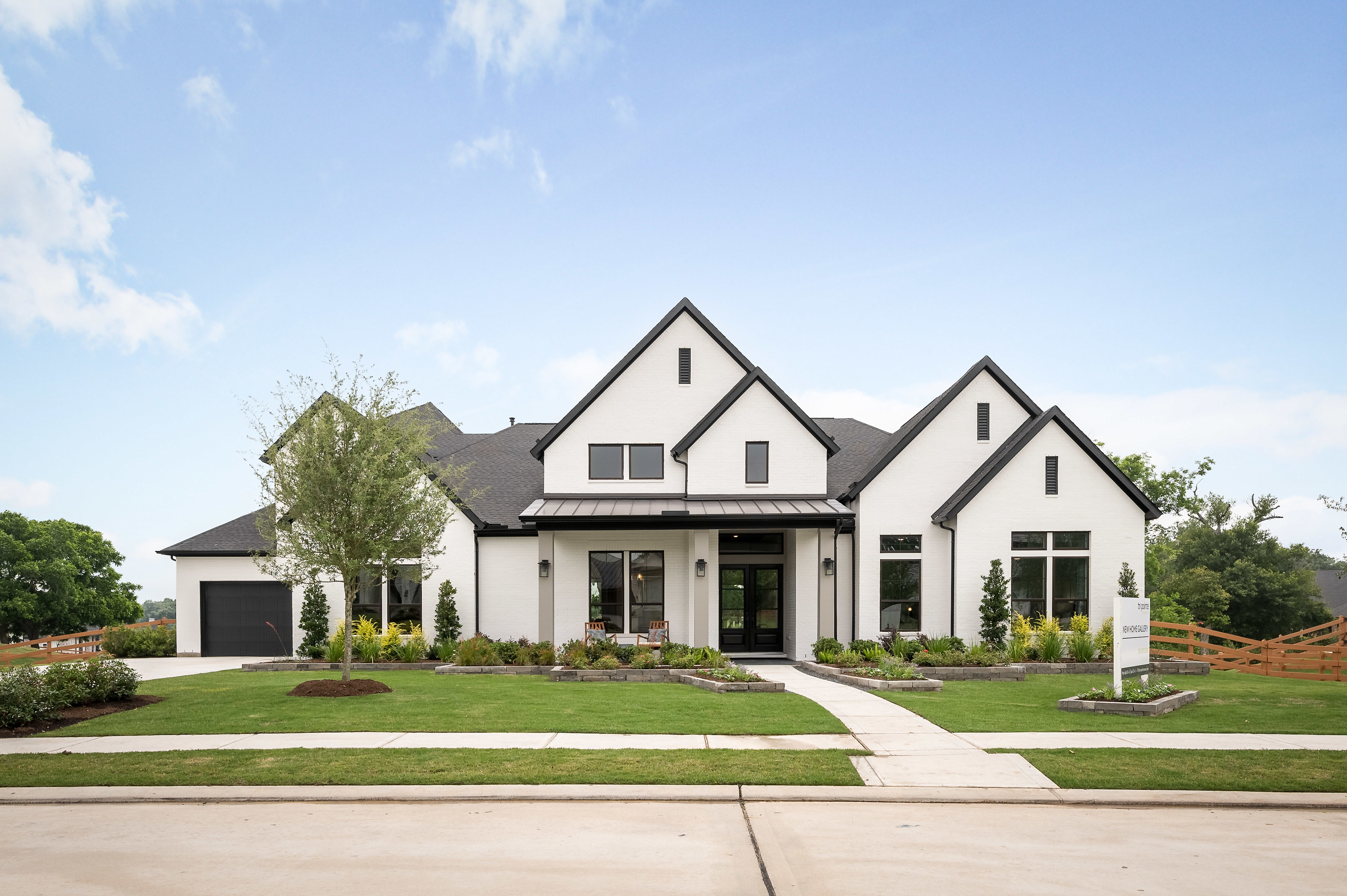 A large, modern farmhouse-style home with a prominent gabled roof and a well-manicured lawn and landscaping in the foreground, set against a clear blue sky with scattered clouds.