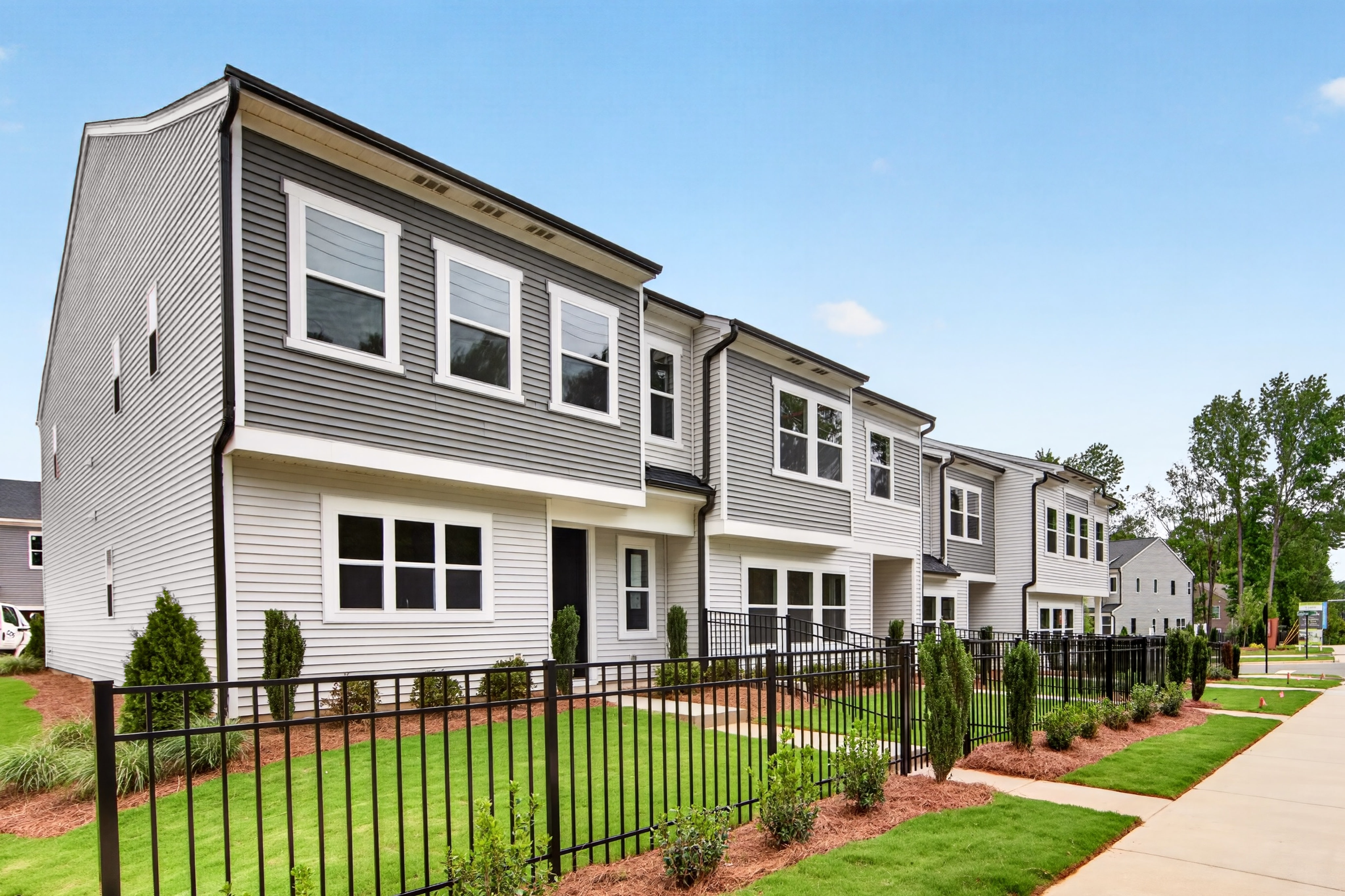 A row of modern townhouses with manicured lawns and black metal fences in the foreground stands against a clear blue sky with scattered trees in the background.