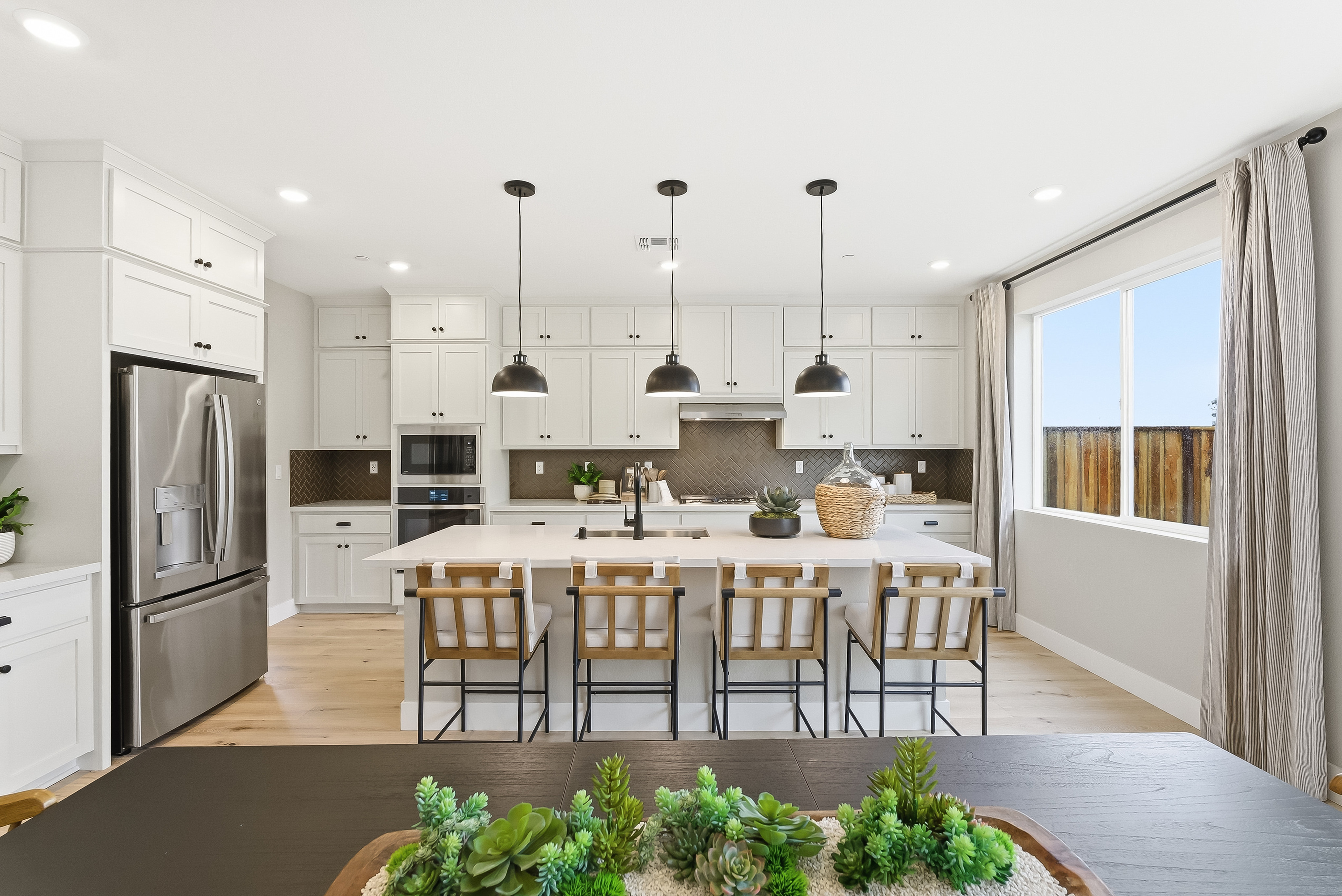 A modern white kitchen with a large island and pendant lights is viewed from a dining table with succulents in the foreground, while a large window reveals a backyard fence in the background.