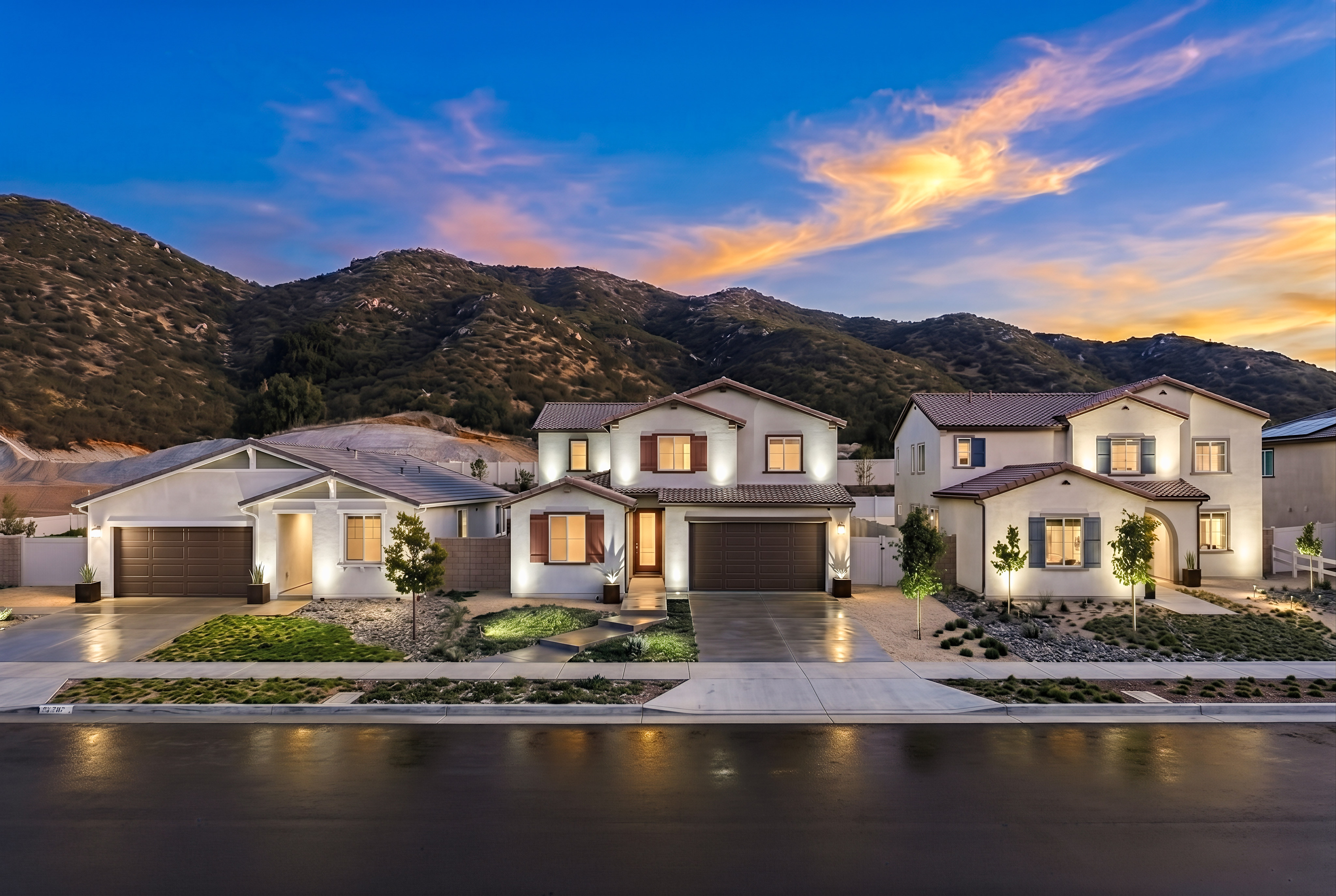 Modern houses with well-lit facades sit in the foreground, while rolling, barren hills under a colorful sunset sky form the background.