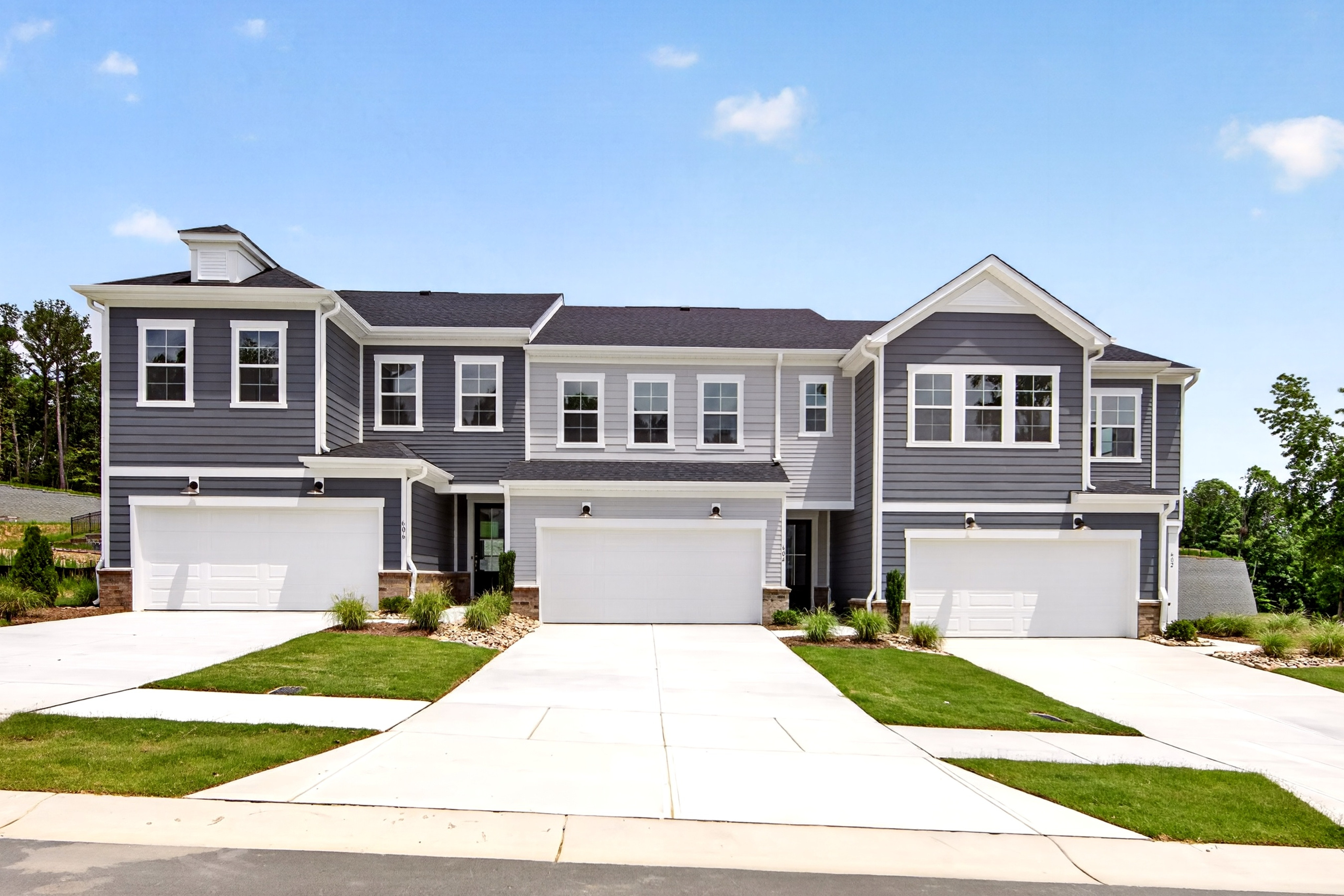 A row of modern townhouses with white garage doors and manicured lawns in the foreground sits against a backdrop of trees and a clear blue sky.