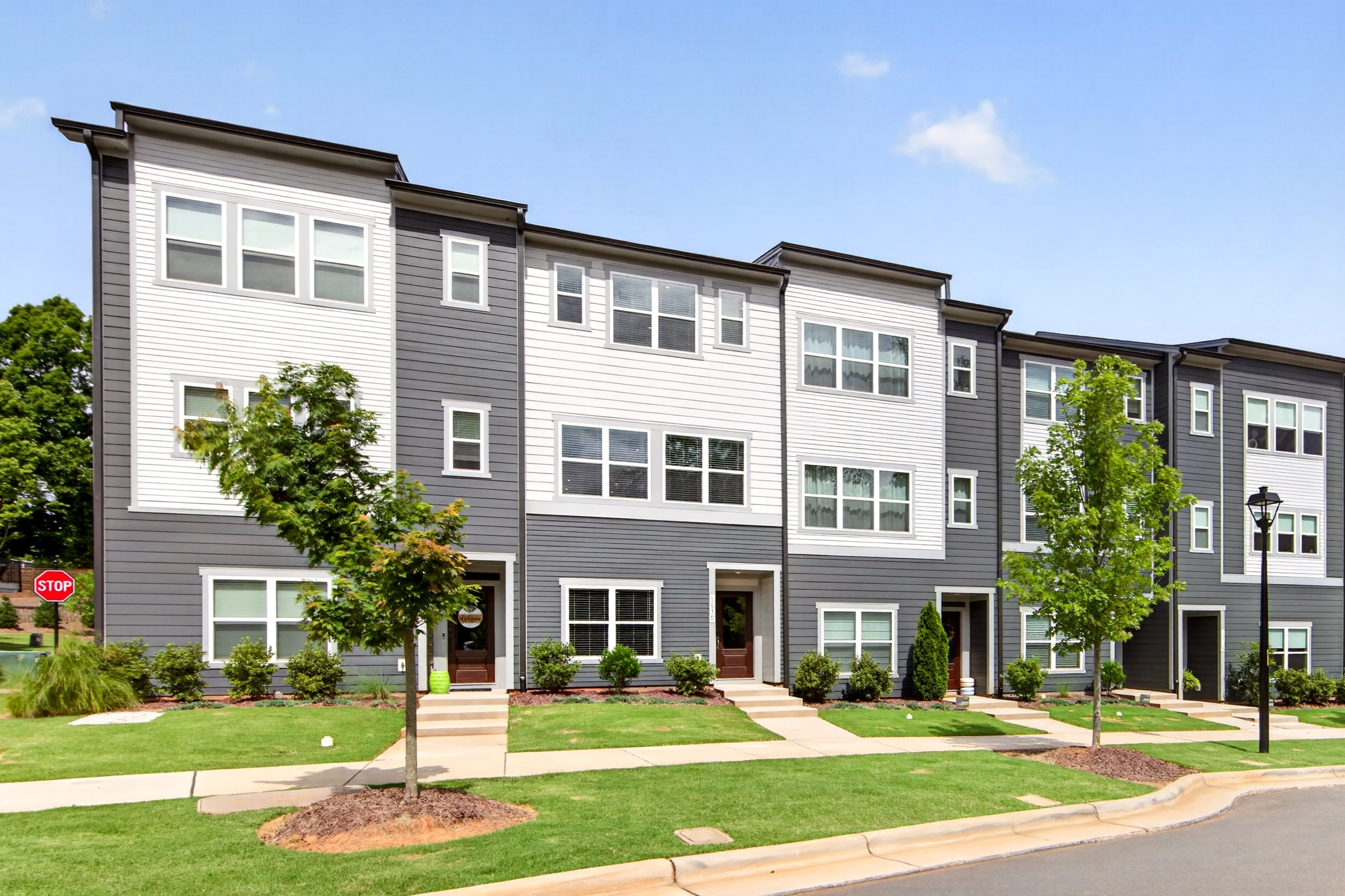 A row of modern townhouses with gray and white exteriors stands on a green lawn with trees in the foreground, against a clear blue sky in the background.