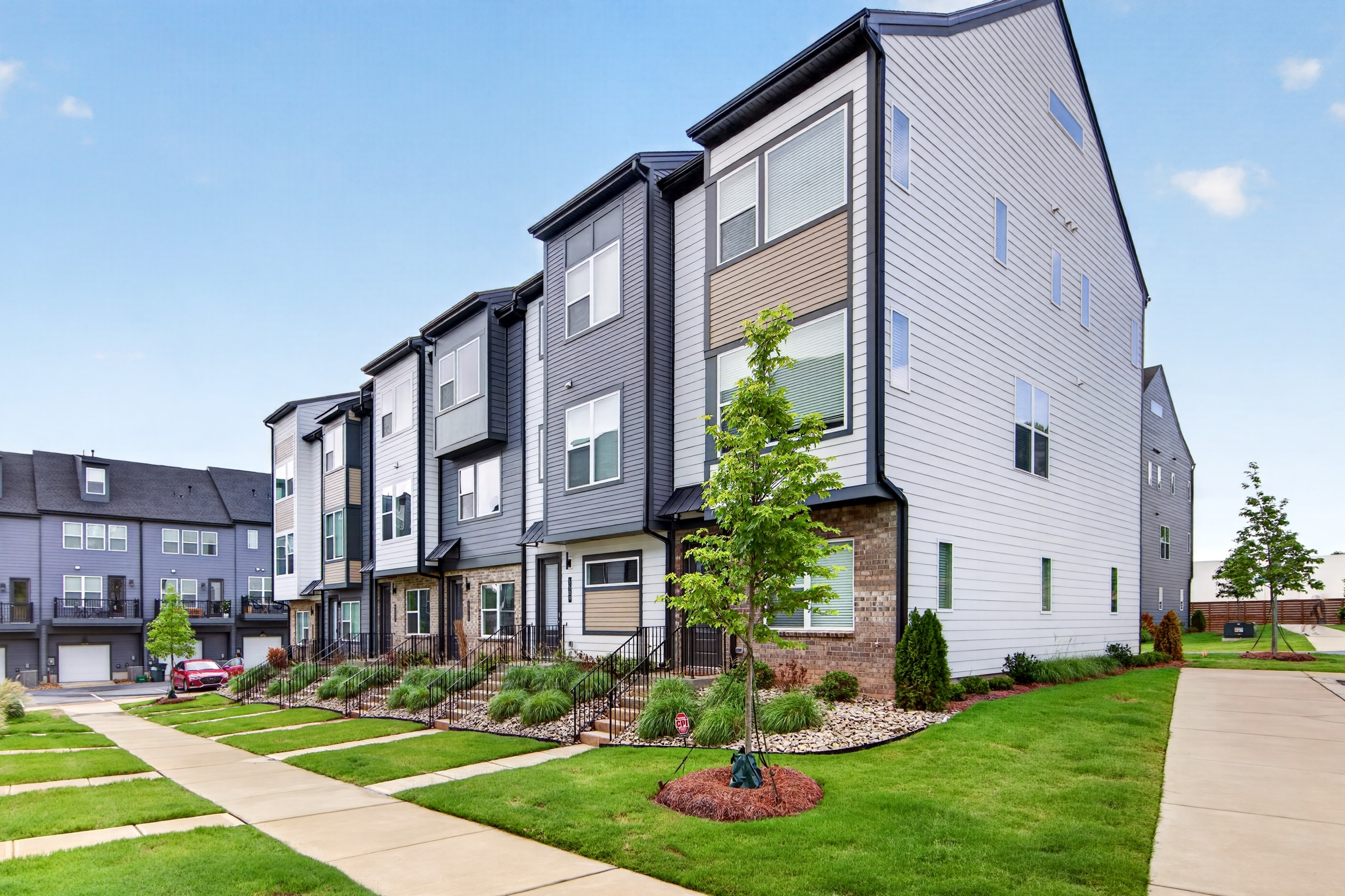 A row of modern townhouses with manicured lawns and a paved walkway in the foreground is set against a clear blue sky in the background.