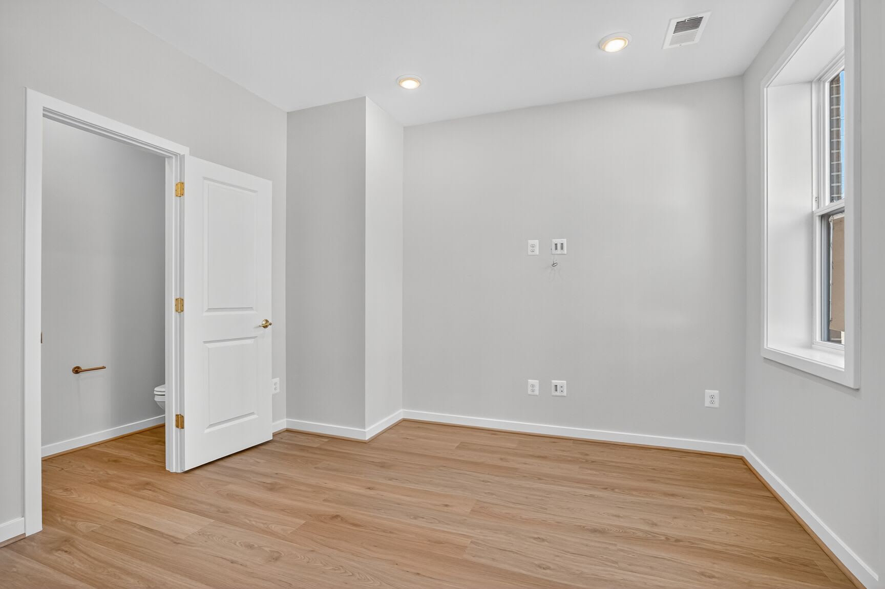 A spacious, well-lit room with white walls, a wooden floor, and a closed door leading to another area.