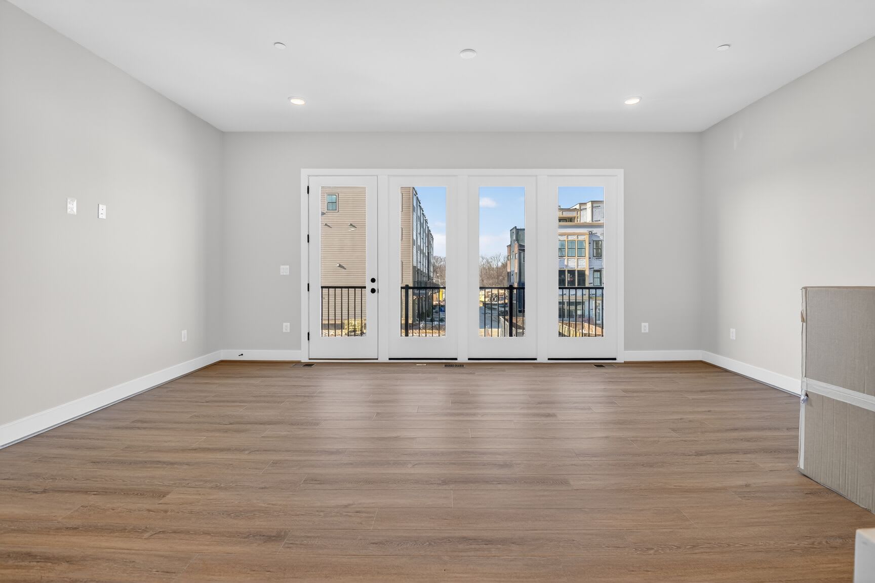A spacious, well-lit room with wooden flooring and large windows overlooking a cityscape in the background.