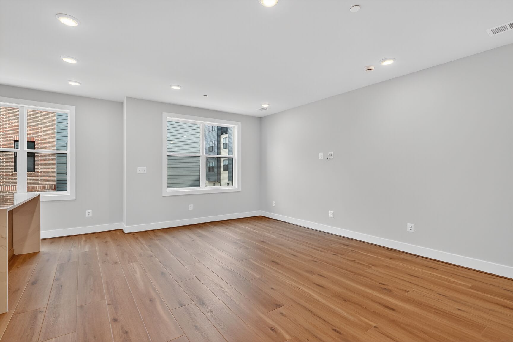 A spacious, well-lit room with hardwood floors, white walls, and recessed lighting fixtures. The room appears to be a living or bedroom space, with a window providing natural light.