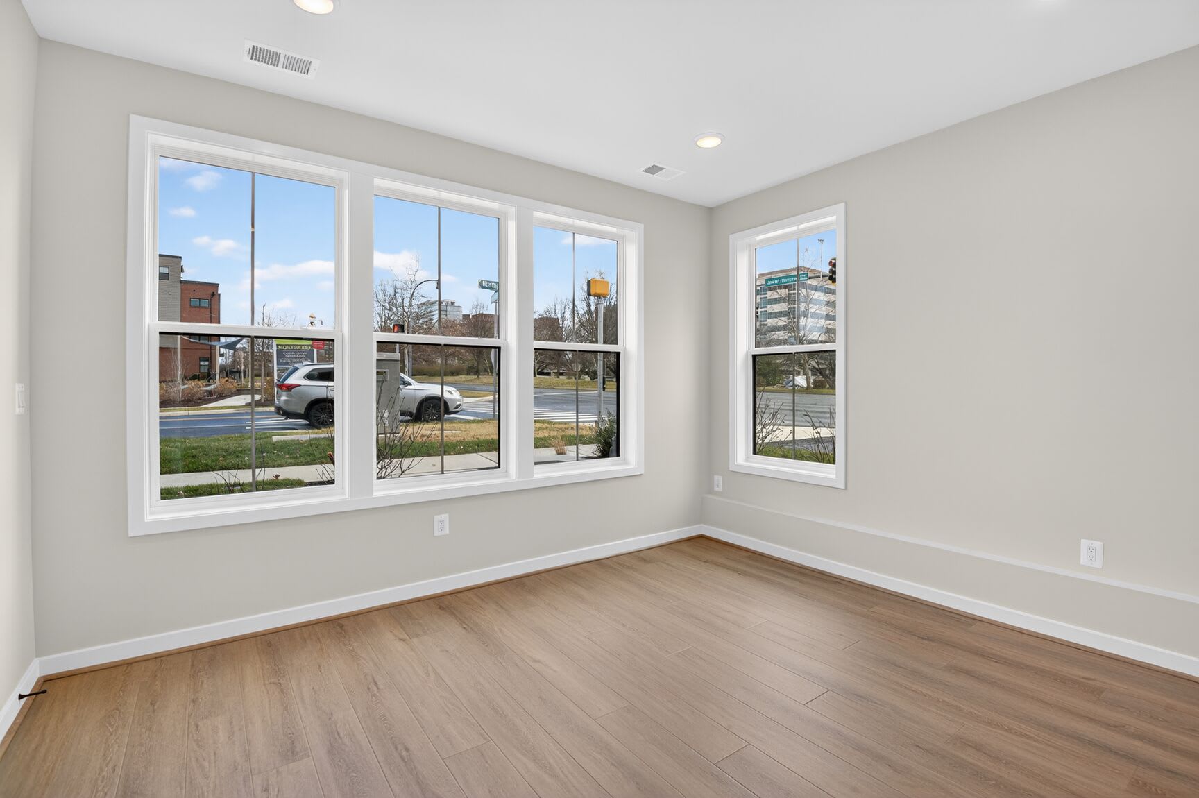 A bright and spacious room with large windows overlooking a residential street, featuring hardwood floors and neutral-colored walls.