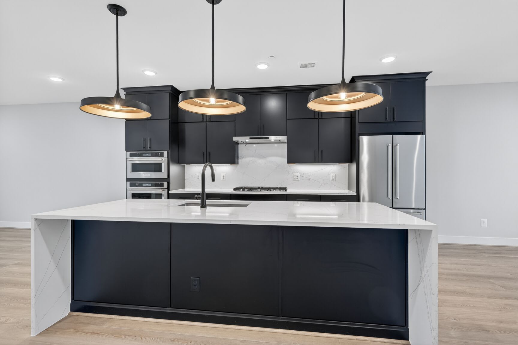 A modern, sleek kitchen with black cabinets, a white island, and three pendant lights illuminating the space.
