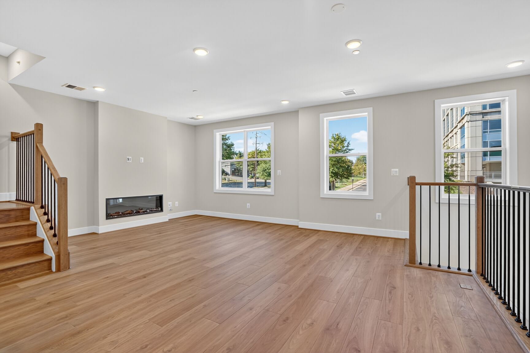A spacious and bright living room with hardwood floors, large windows, and a fireplace set against a neutral-toned wall.