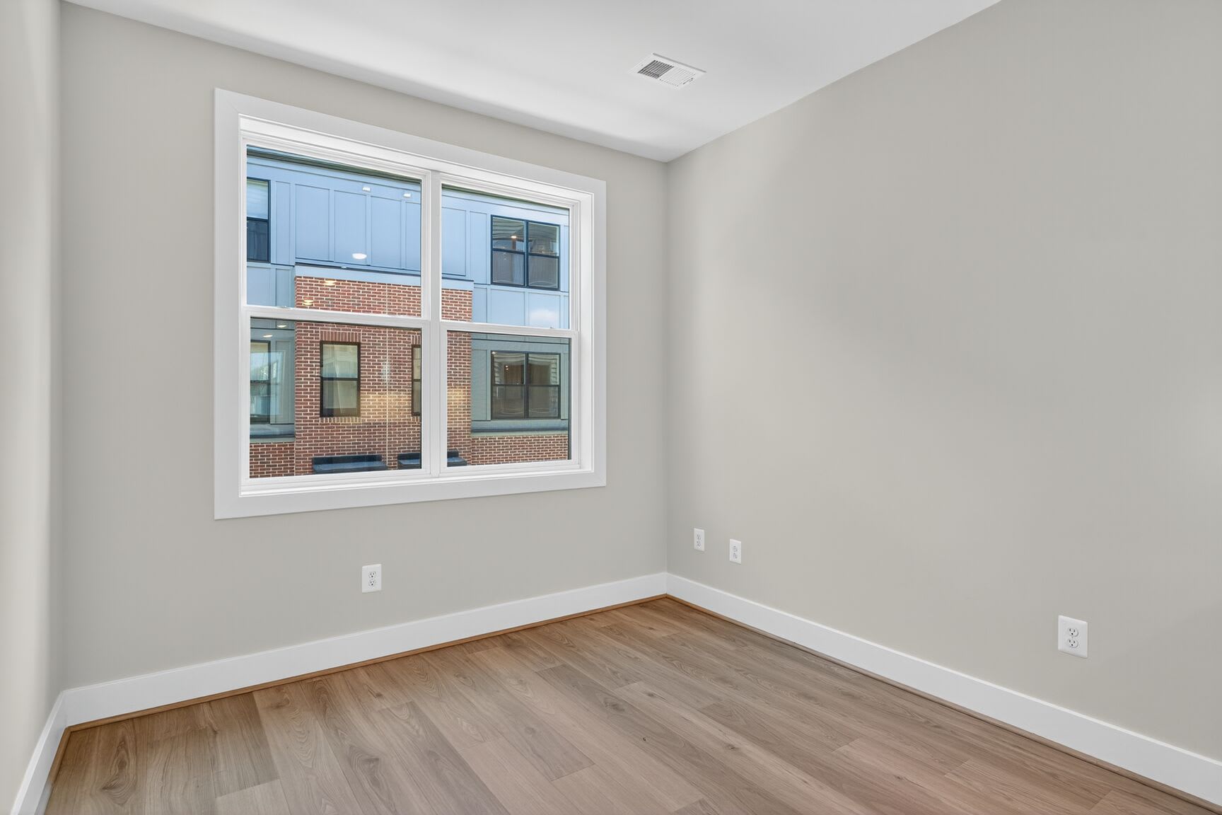 A spacious room with a large window overlooking a brick building, featuring hardwood floors and neutral-colored walls.